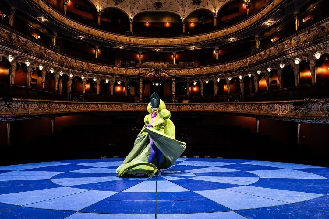 Seth Carico performs in a bright green flowing costume, alone on a circular blue stage with ornate balconies for the audience.
