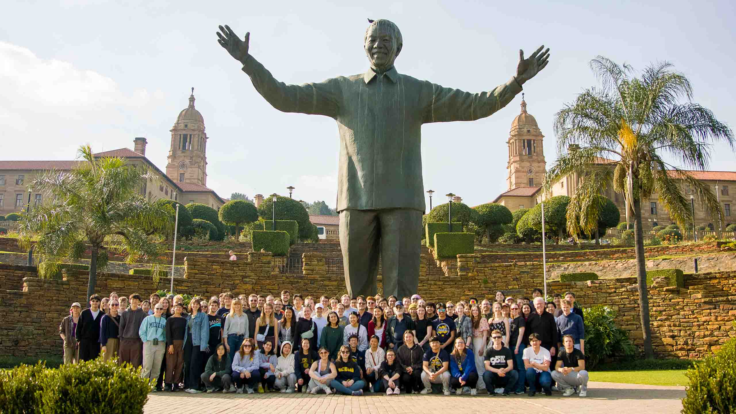 The USO poses beneath a statue of Nelson Mandela. His statue is green, very tall, and he outstretches his hands in the air.