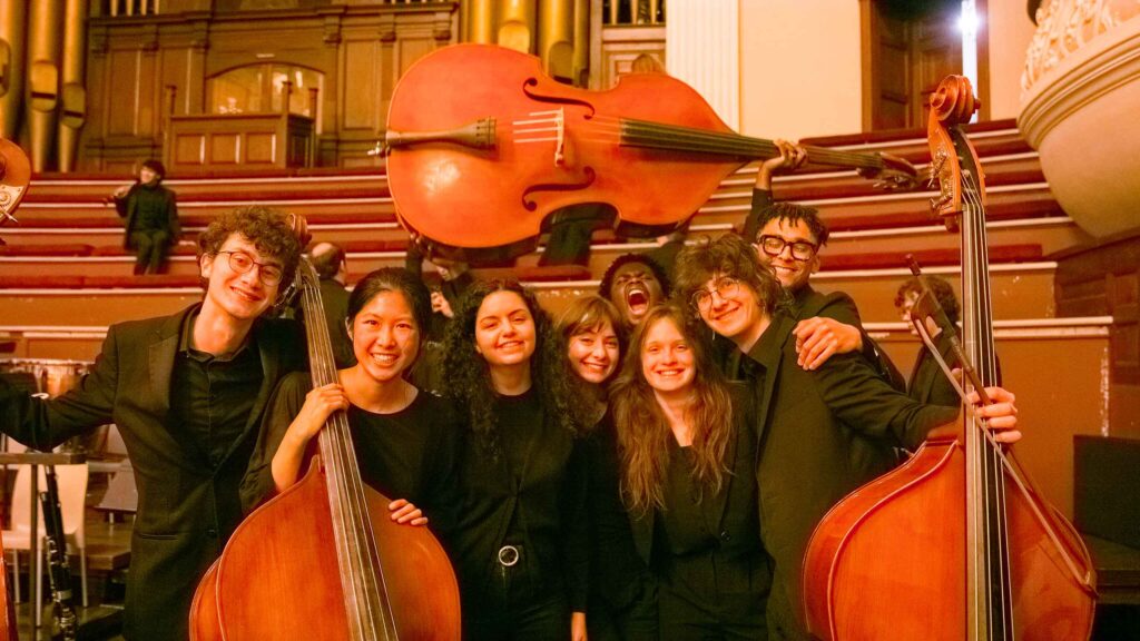 A group of 8 students poses with 3 double basses, attired in concert black.
