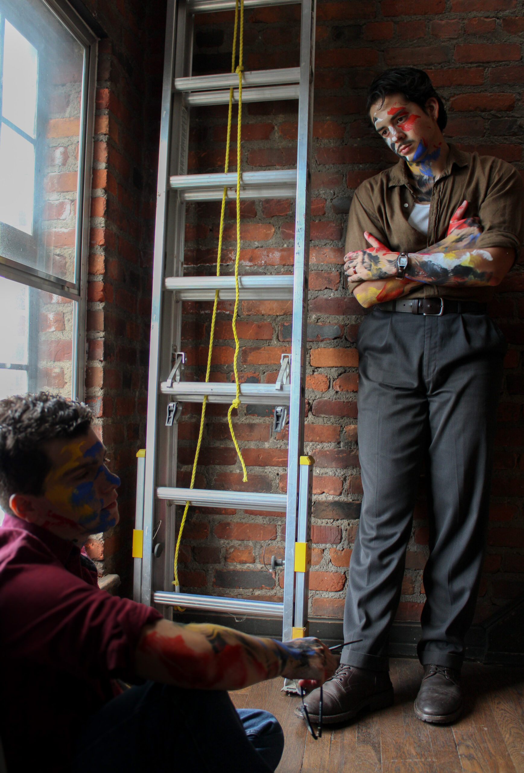 Two actors, one standing and one sitting, both covered in paint near a ladder by a window.