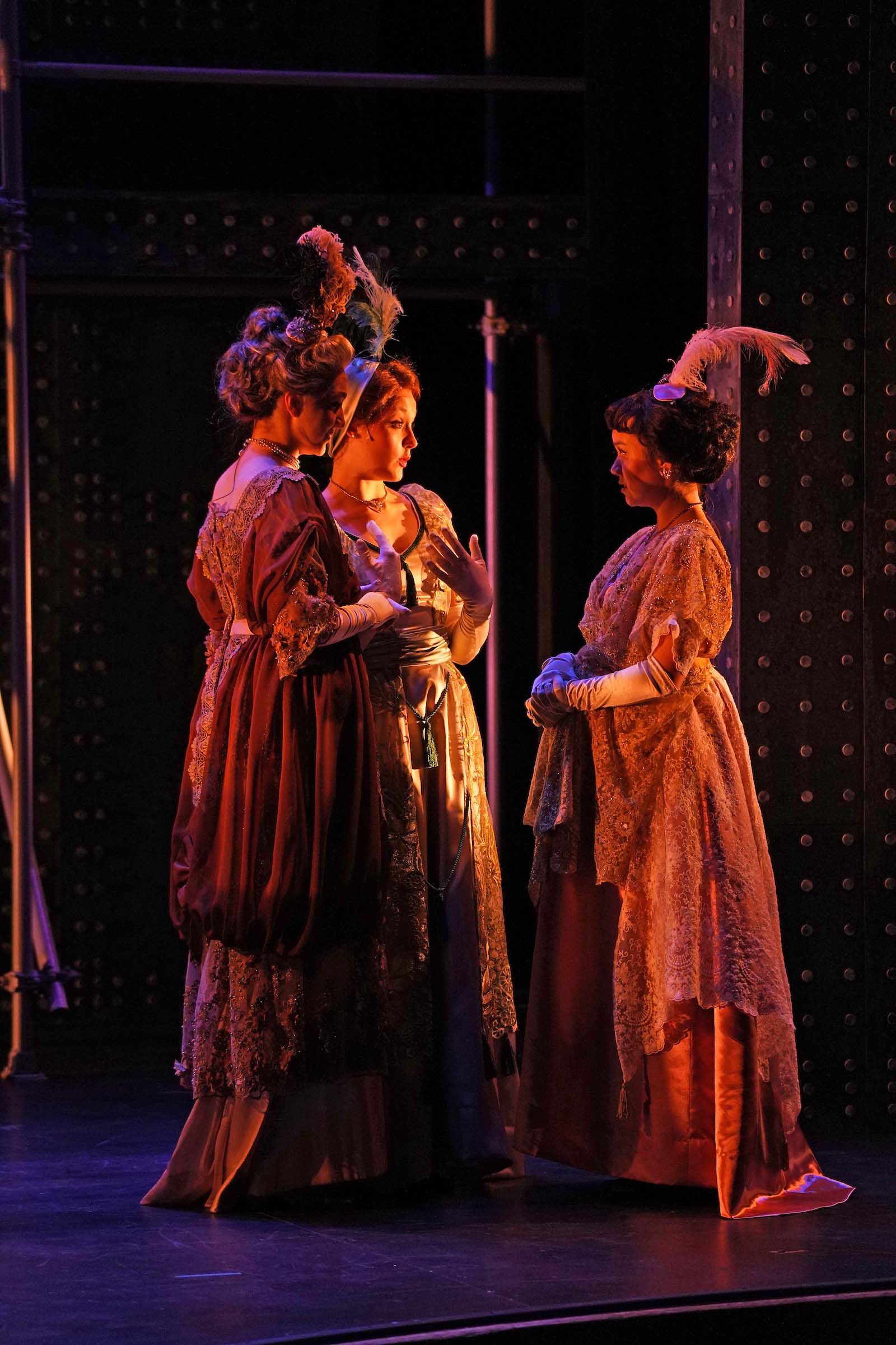 2d Mrs Astor Titanic Emily Murakami completed costume Three actors perform standing on stage in Titanic: The Musical, attired in evening gown costumes with feather headpieces.