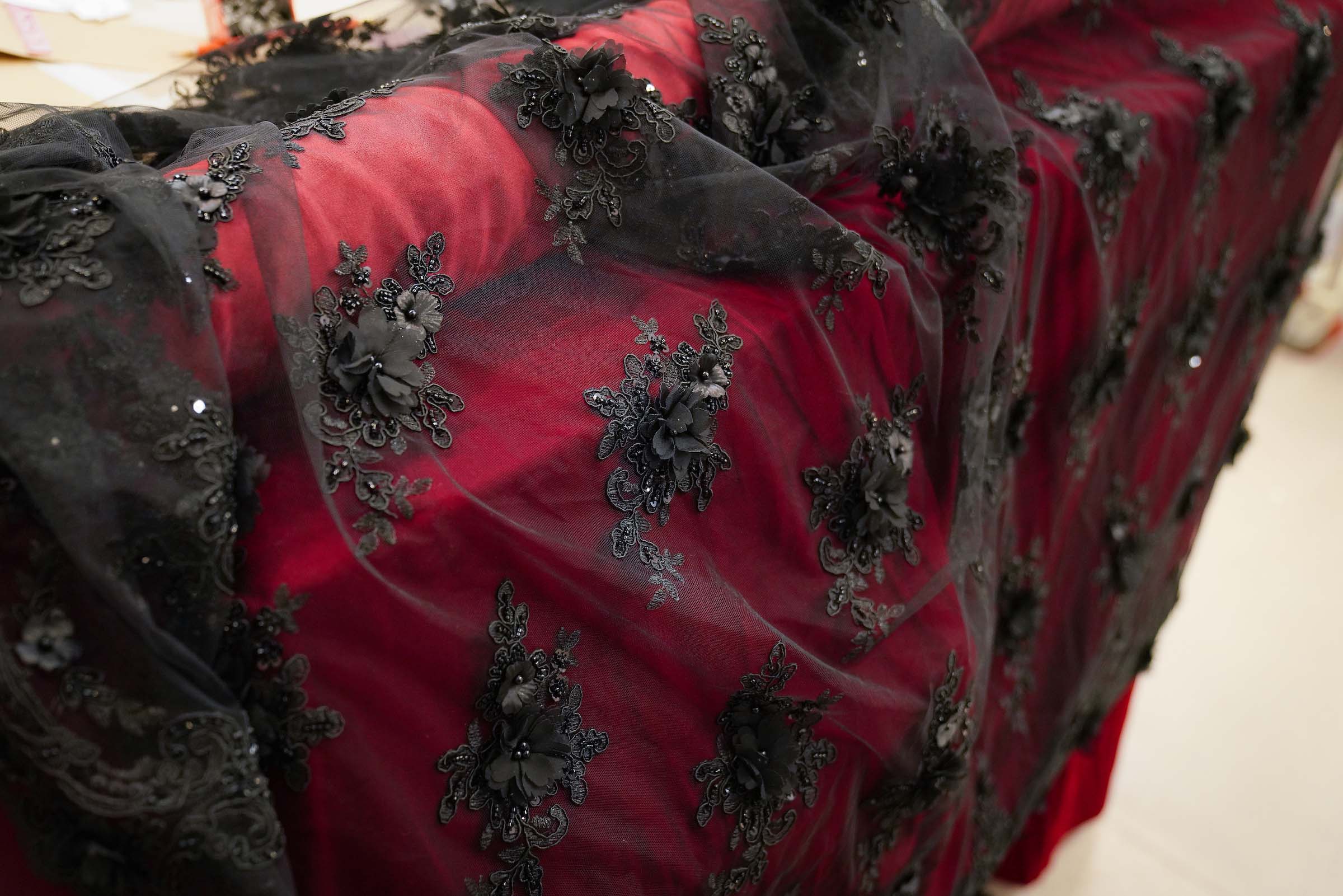 4a fabric Costume Shop Titanic A roll of deep red satin fabric, covered with a sheer black overlay fabric embroidered with a pattern of black flowers with beads.