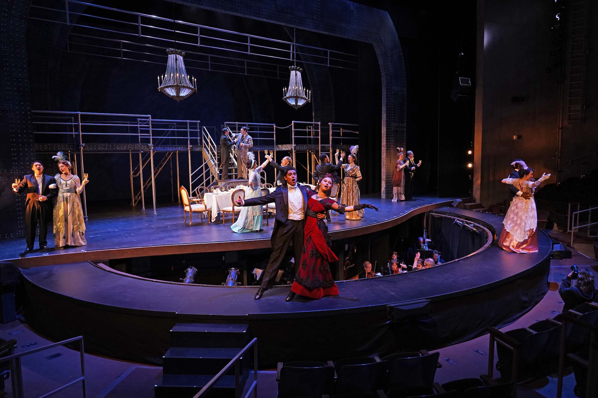4b evening dress Costume Shop Titanic At the Power Center, the cast and orchestra for Titanic: The Musical perform; at center stage, a couple dances together, including a performer costumed in a red satin evening gown with sheer black embroidered overlays.
