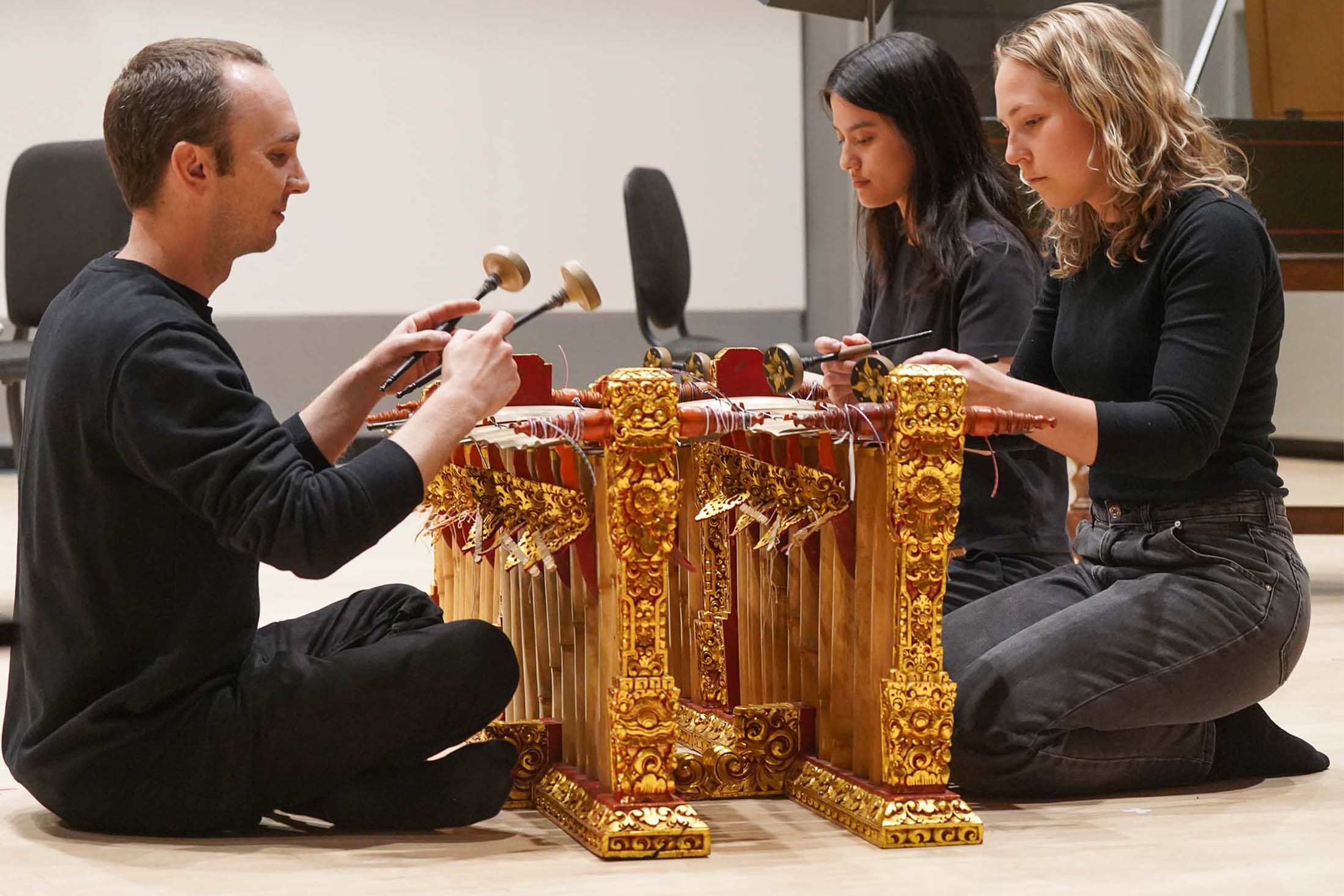 Four students in the Gamelan Ensemble perform using percussion mallets, seated on the floor.