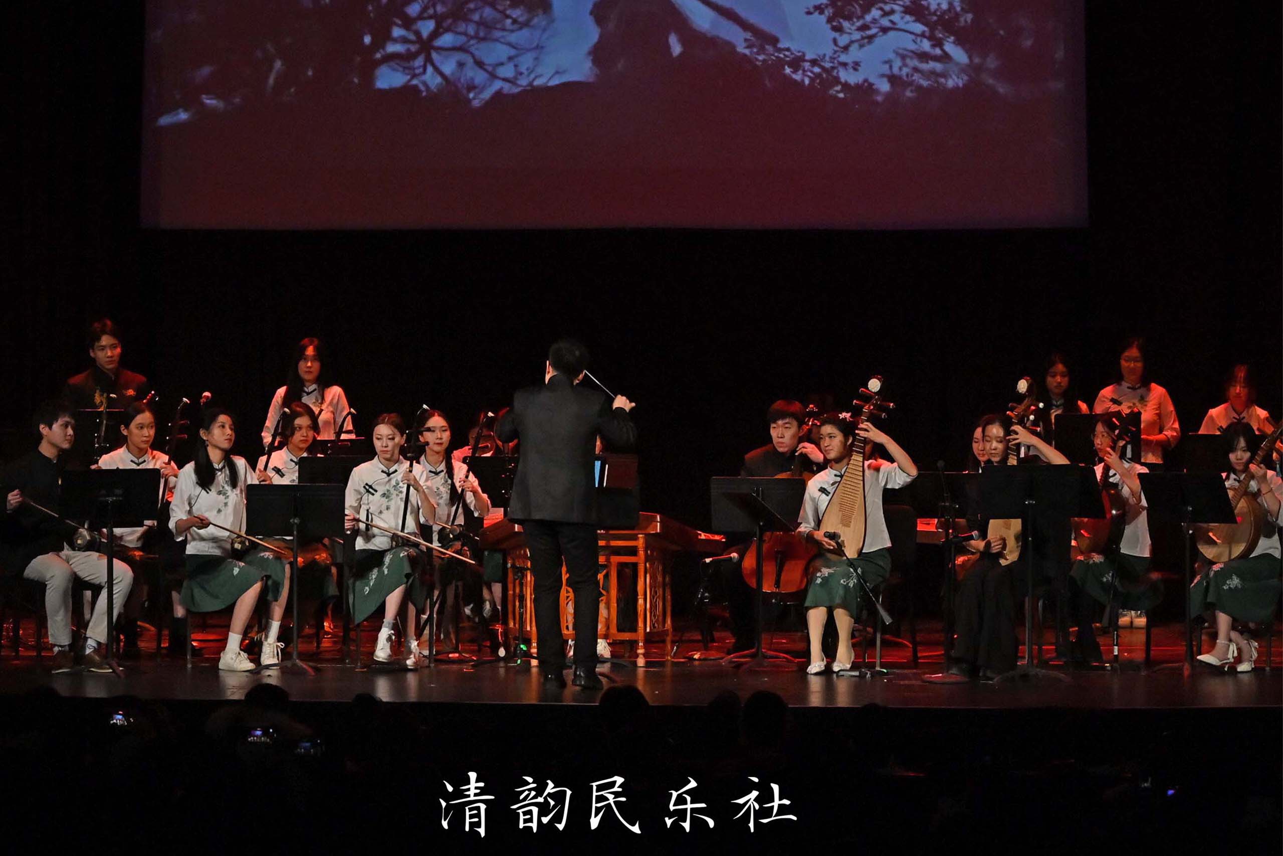 The Qingyun Chinese Instrument Ensemble performs seated on a stage, with a projection displayed above.