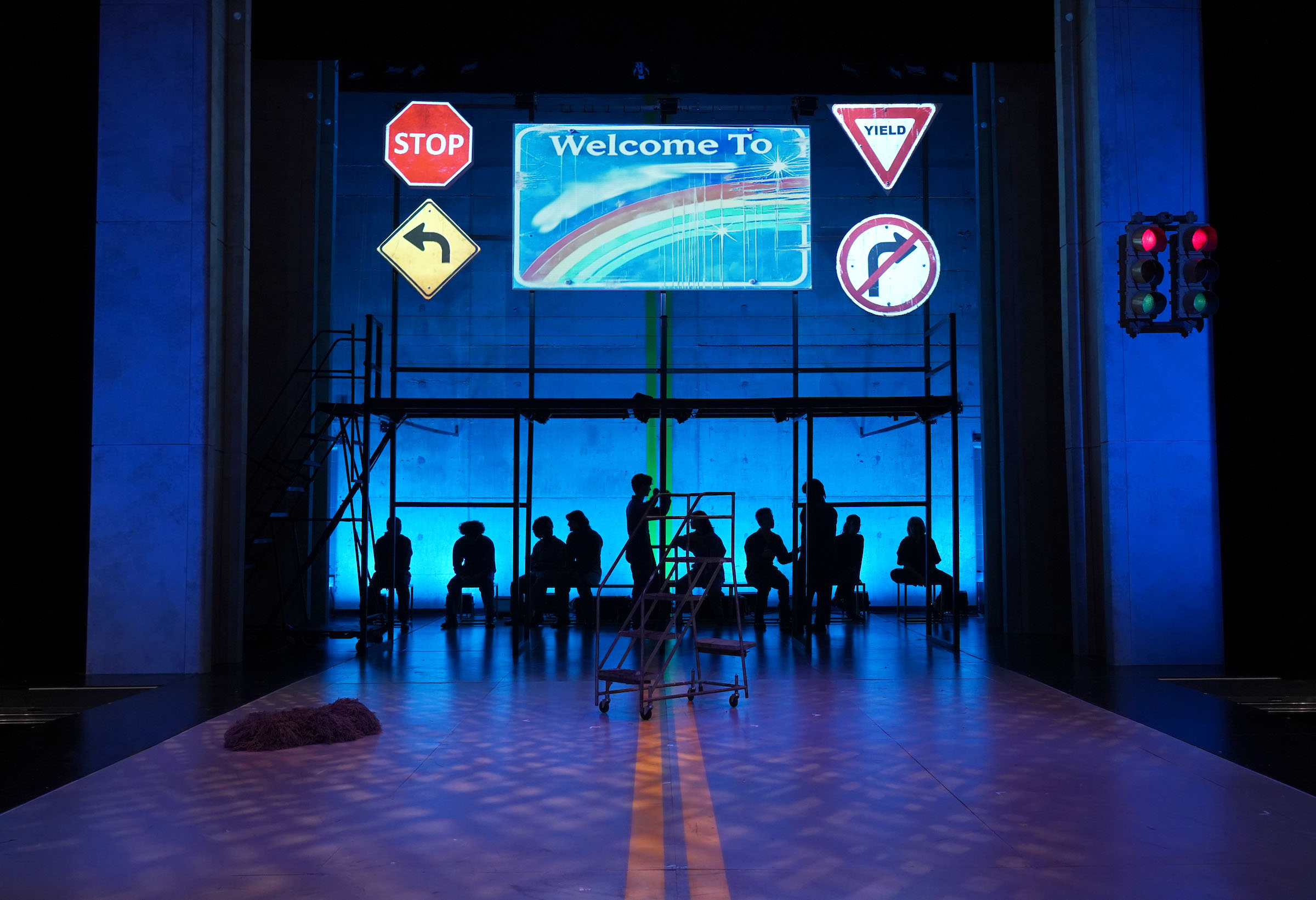 Theatrical Shops Overview Our Oz Arthur Miller Theatre 2025 Stage scenery lit in blue-tones and bright street and traffic signs; one shows the words "Welcome to" and a rainbow. Below, we see shadow figures of the cast.