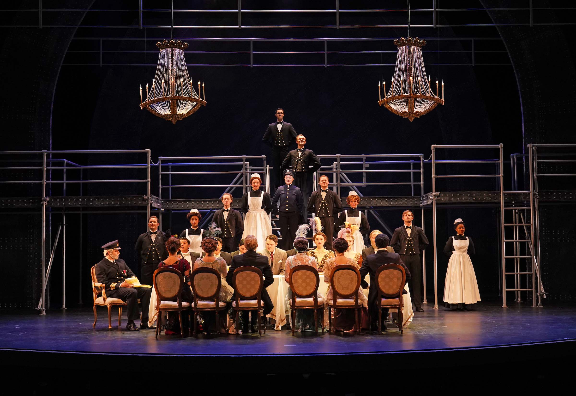 On the stage set of Titanic: The Musical, First Class passengers dine while crew members look on, with crystal chandeliers hanging above.