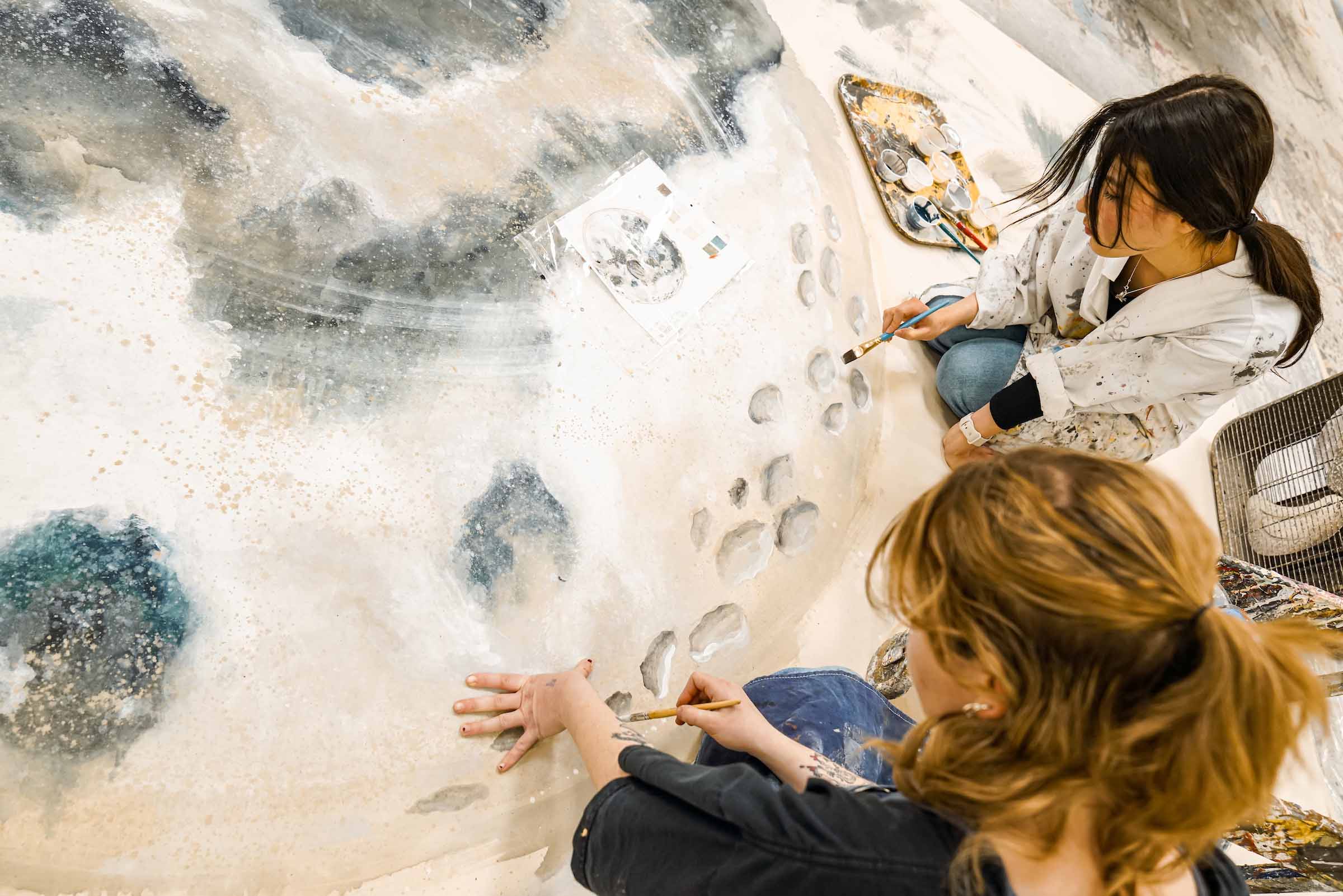 Two students kneel down while painting moon details on a large canvas on the floor of the shop.