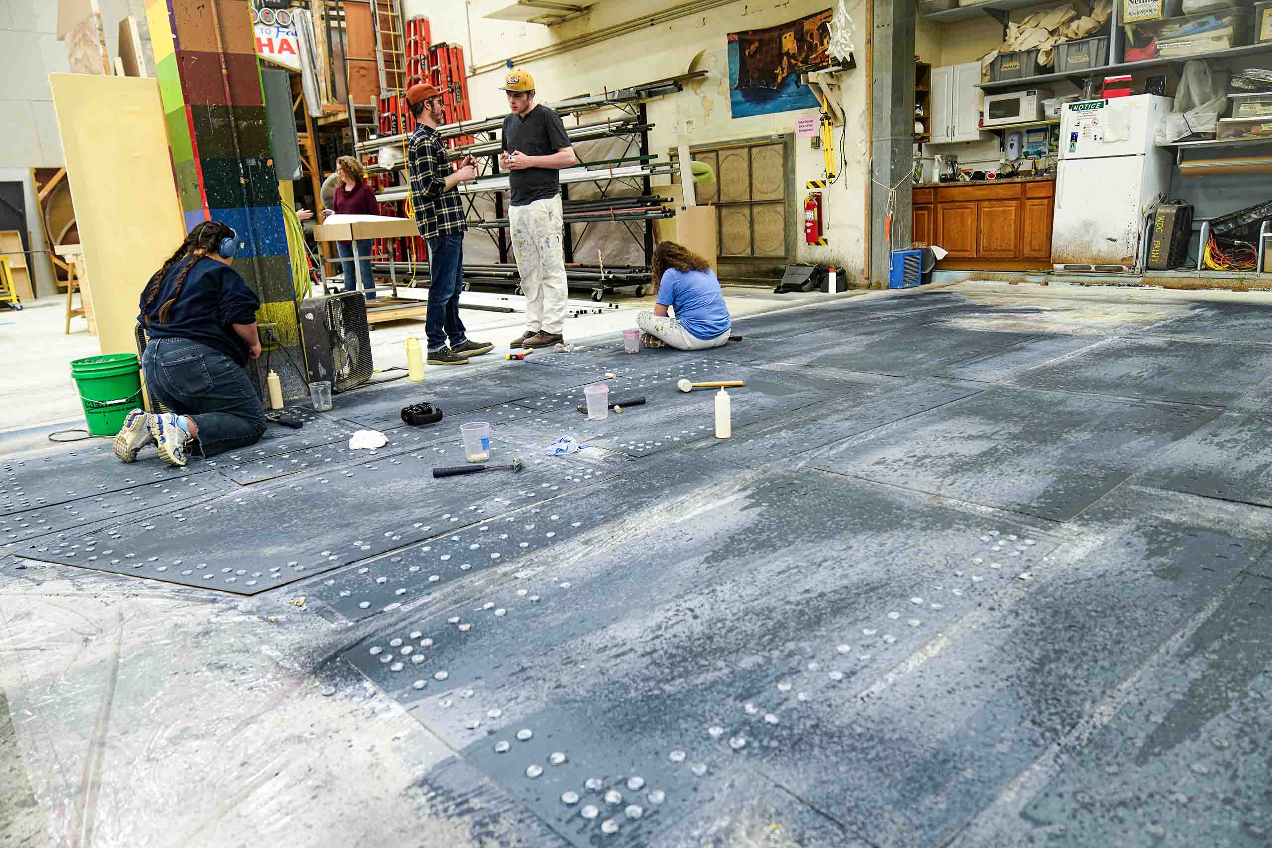 2a Scene Shop students rivets ship Titanic In the scene shop, huge panels painted like grey steel are laid out on the floor, and two students kneel and sit on them while adding rivets.