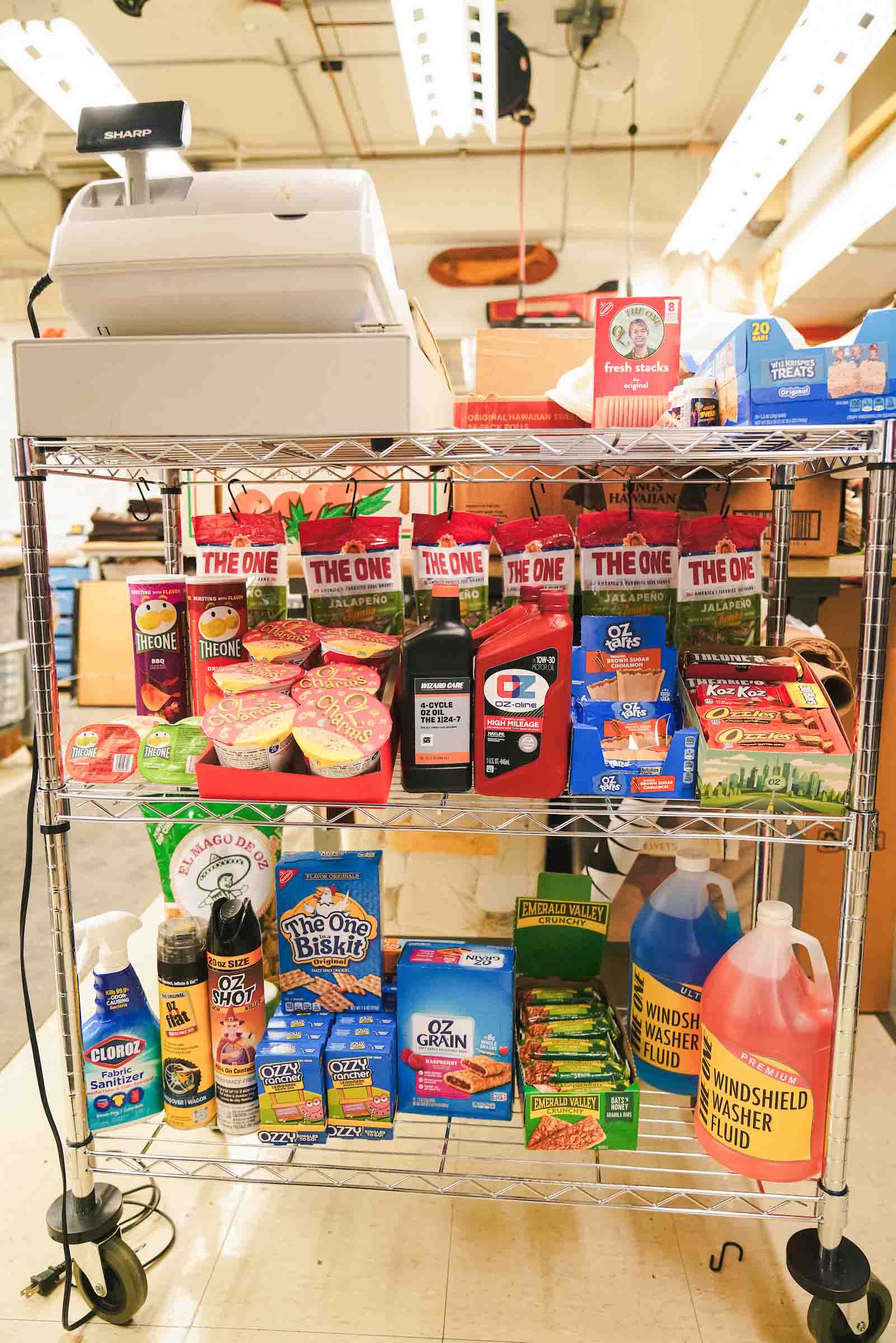 A wheeled set of assembled aluminum shelves, lined with Oz- themed packaged grocery items and a cash register on top.