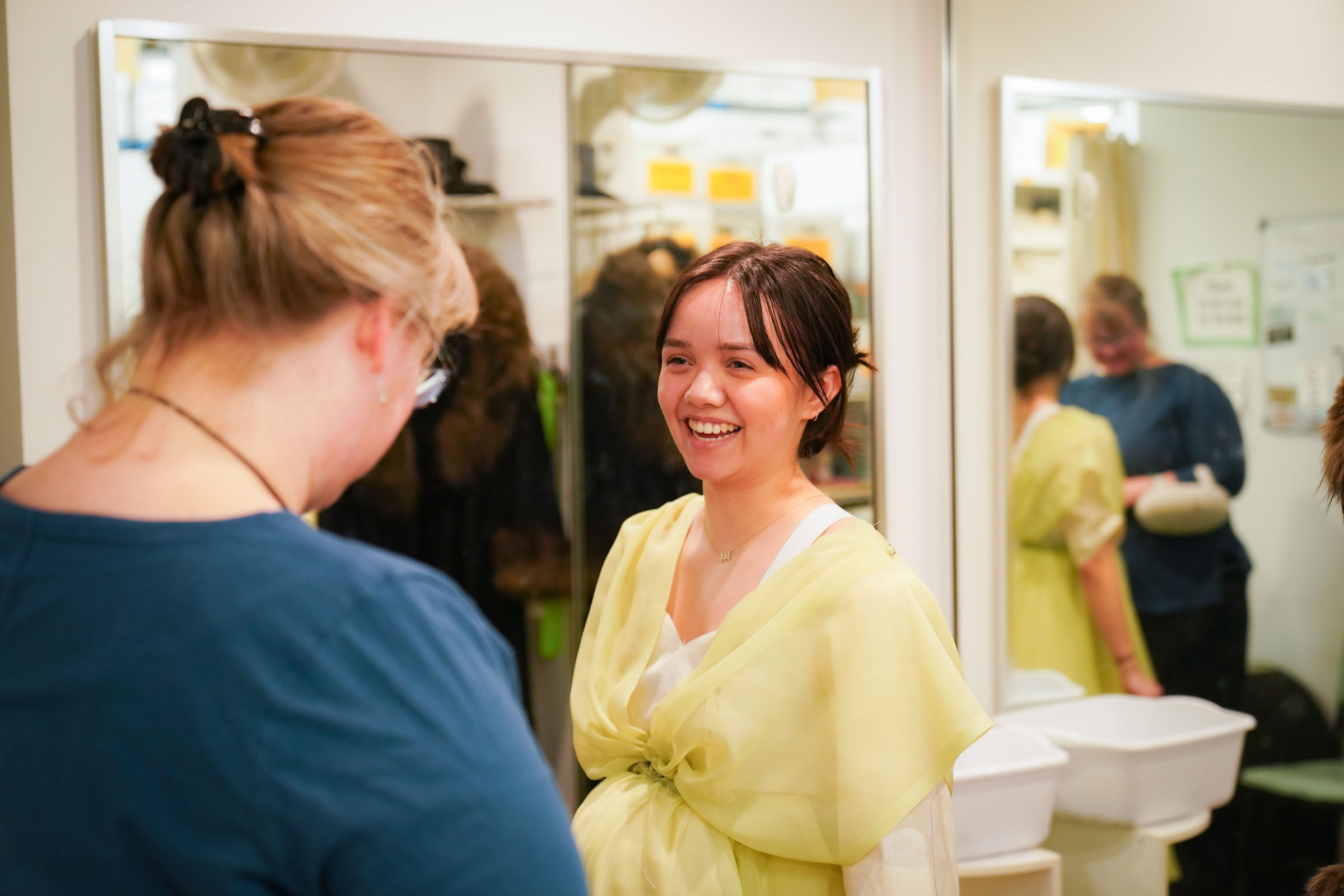 2b Leslie Ann Smith with Emily Murakami costume fitting Mrs Astor Titanic Emily Murakami smiles at Leslie Ann Smith, wearing an unfinished costume made of yellow fabric, in a space lined with mirrors.