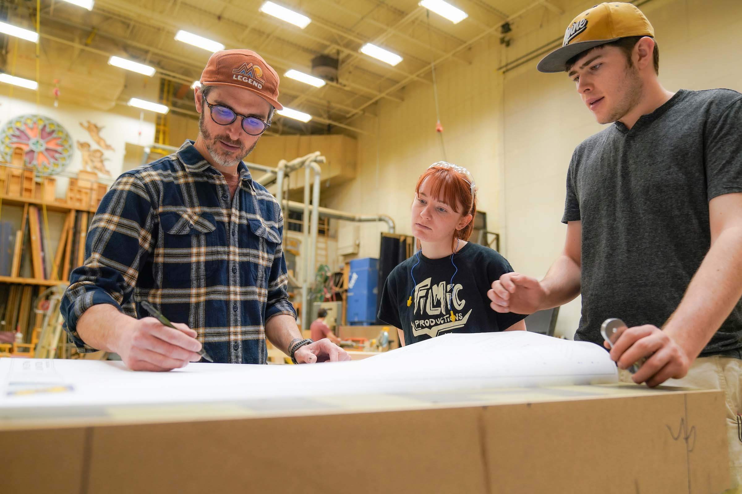 3 Chad Hain with students Power Center Scene Shop In the scene shop, three people work together standing at a table, focused on a large drafting document.