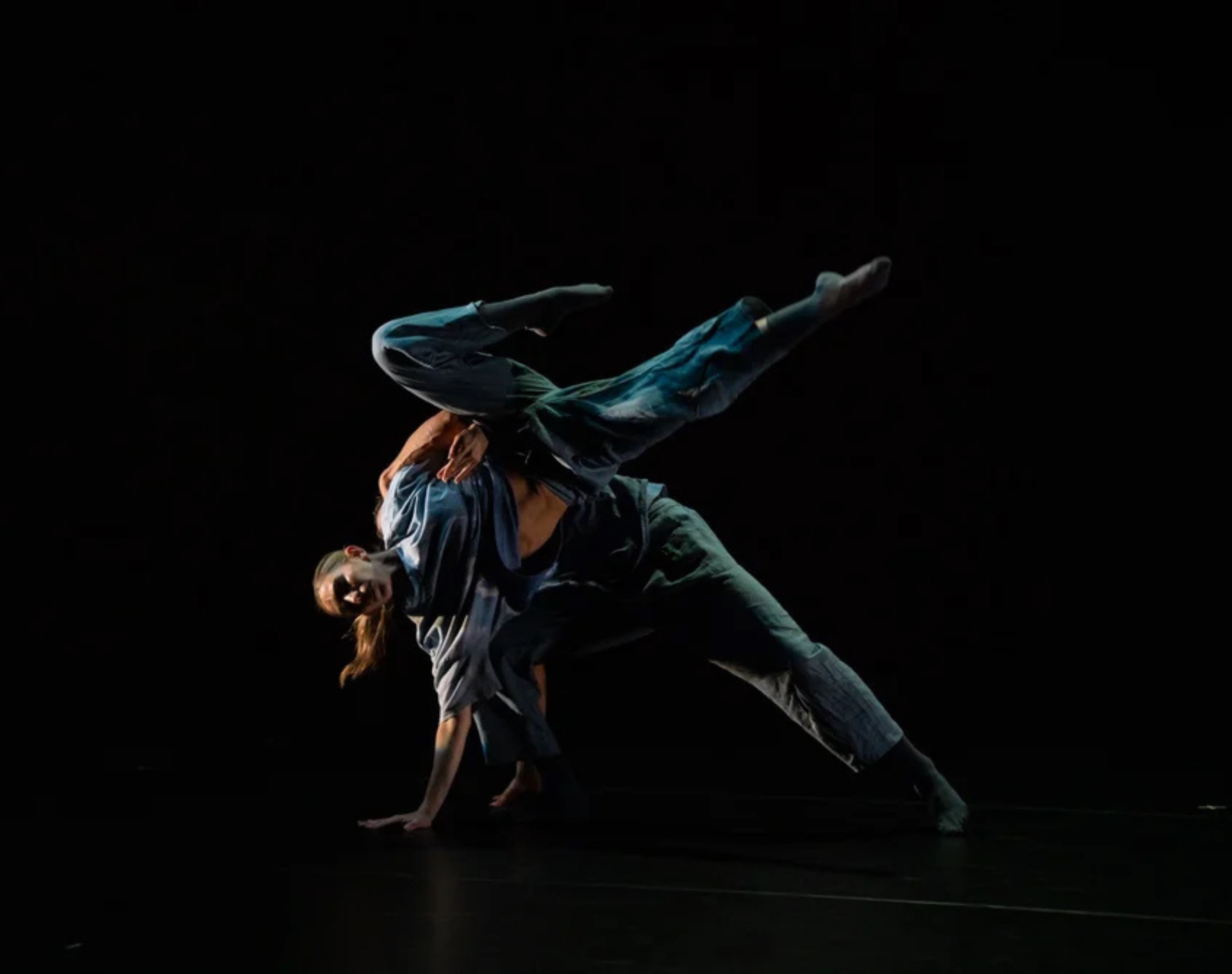 Two dancers from Eisenhower Dance Detroit, wearing loose, blue clothes, dance together against a black background