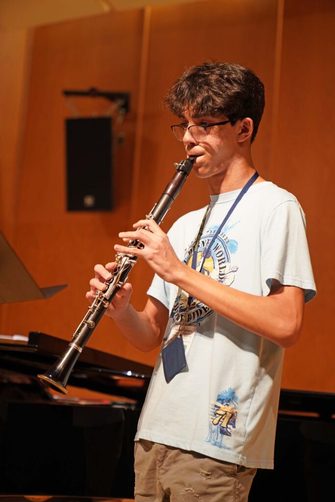 An MPulse student plays clarinet standing on stage, wearing summer attire and a hanging name tag.