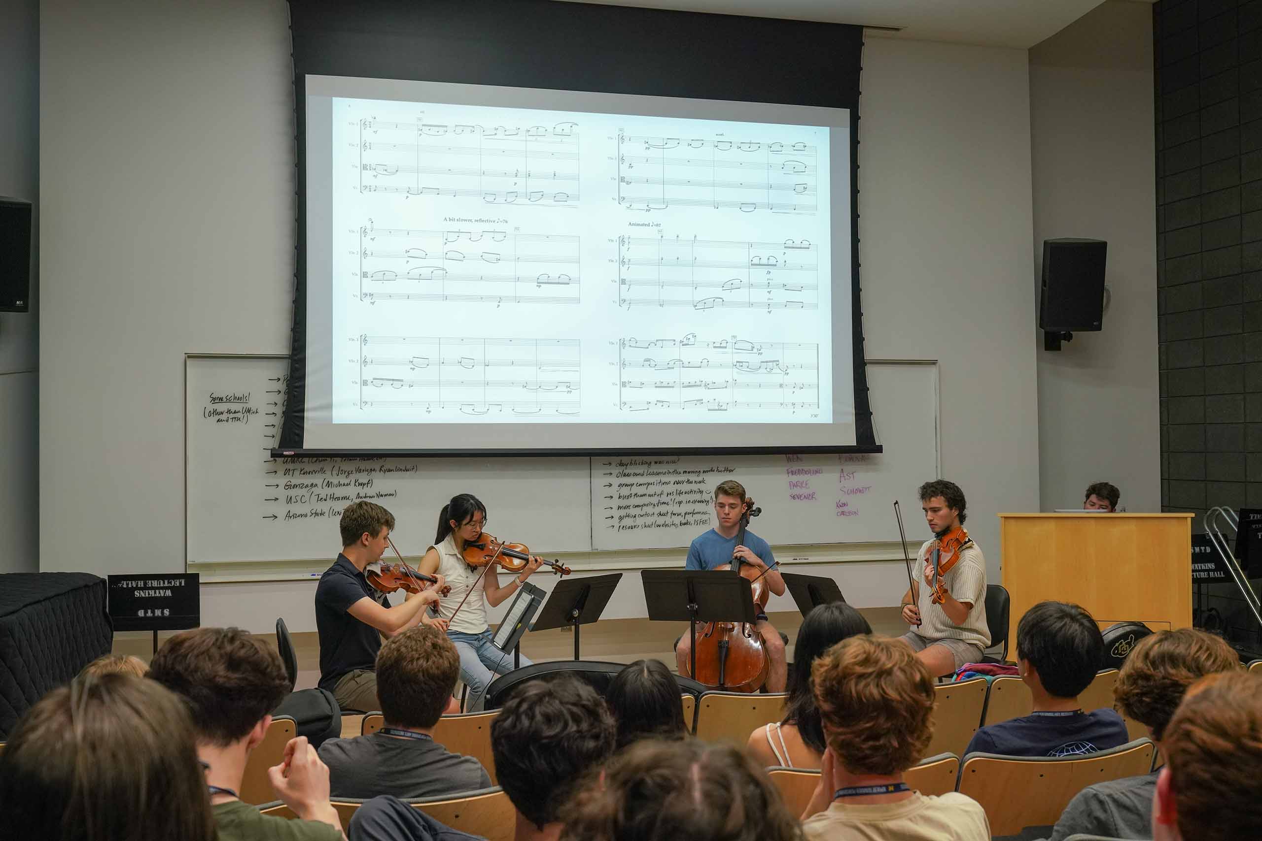 A string quartet performs for a group of students. A screen of the sheet music is projected on top of the musicians