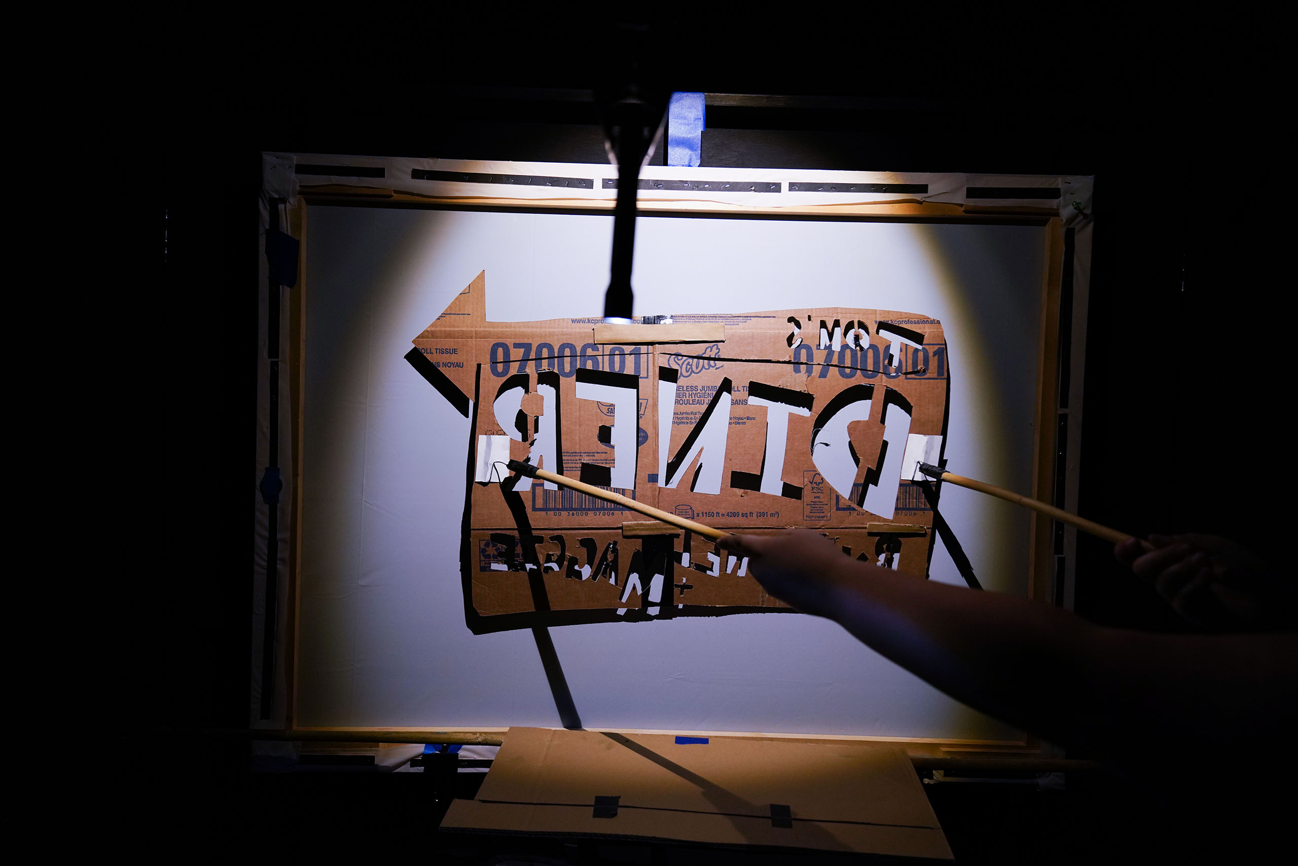 A spotlight shines on a cardboard shadow cut-out sign for TOM'S DINER, as part of a theatrical presentation.