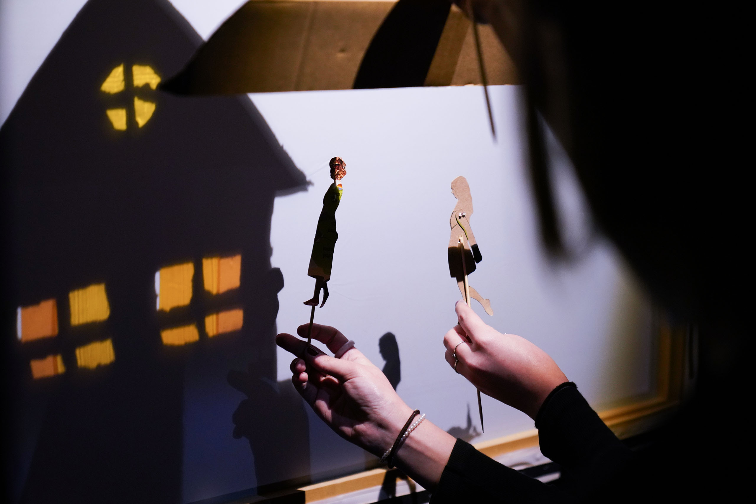 Two people hold up and move shadow puppets next to a house in a theatrical presentation.