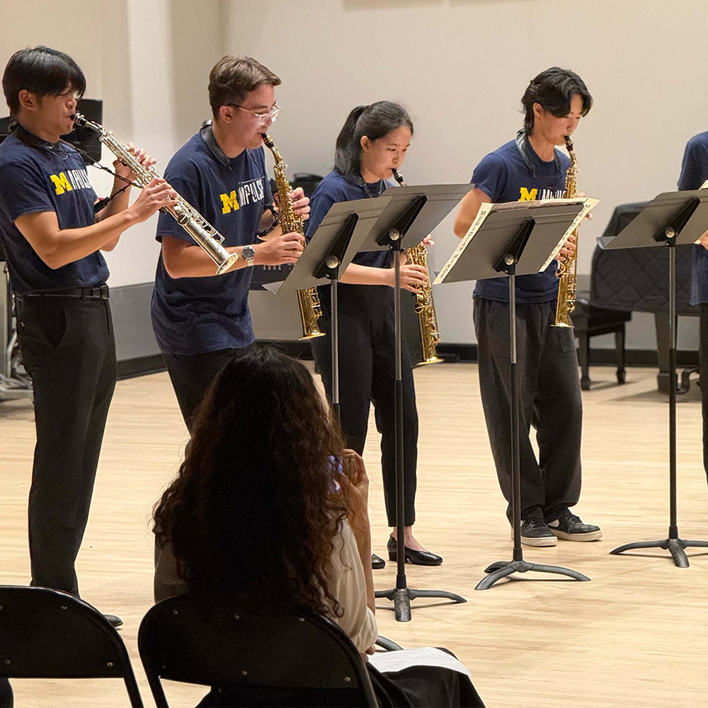 Four MPulse students playing soprano saxophones perform standing, with an audience member seated in the foreground.