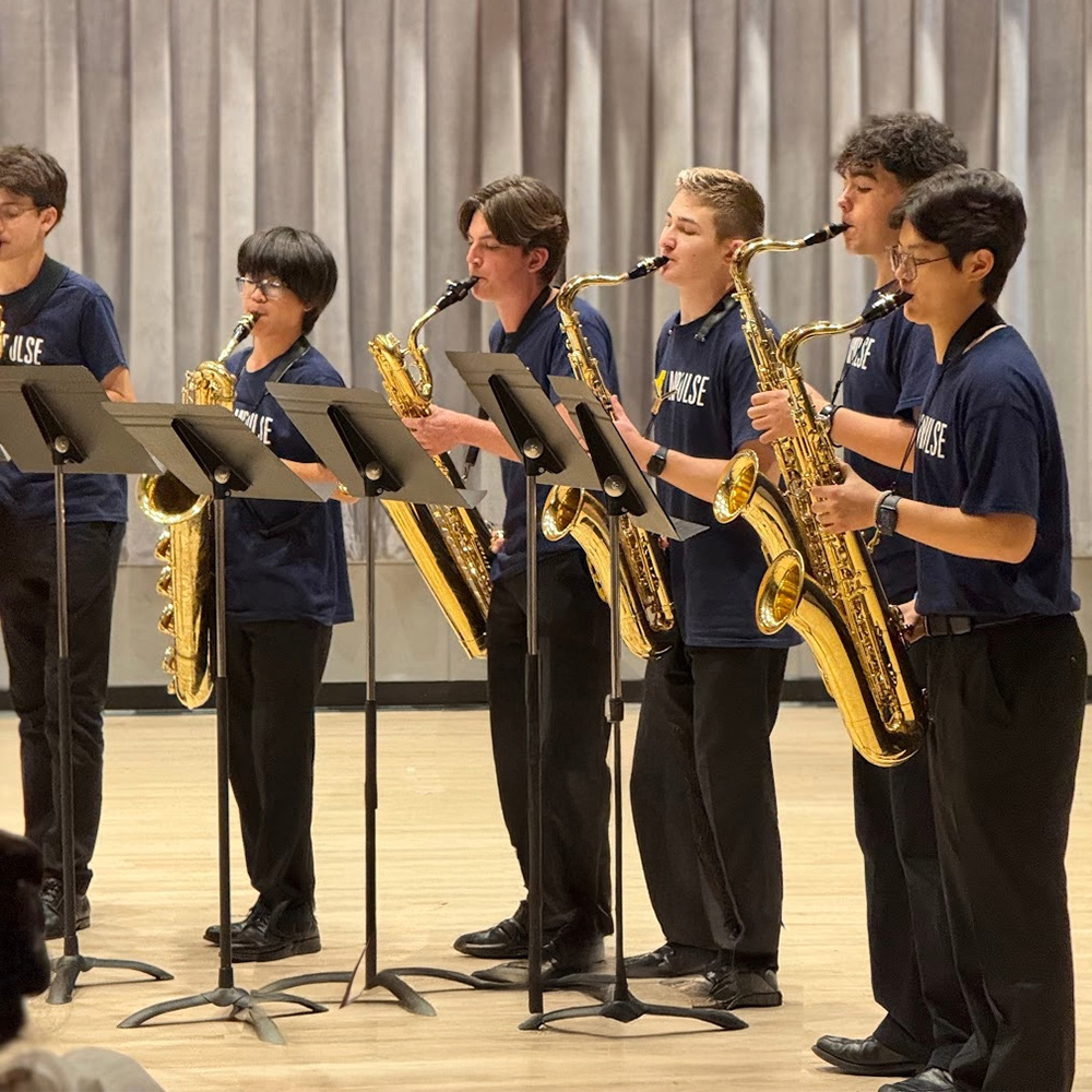 Six MPulse students perform standing with music stands, playing tenor and bass saxophones.