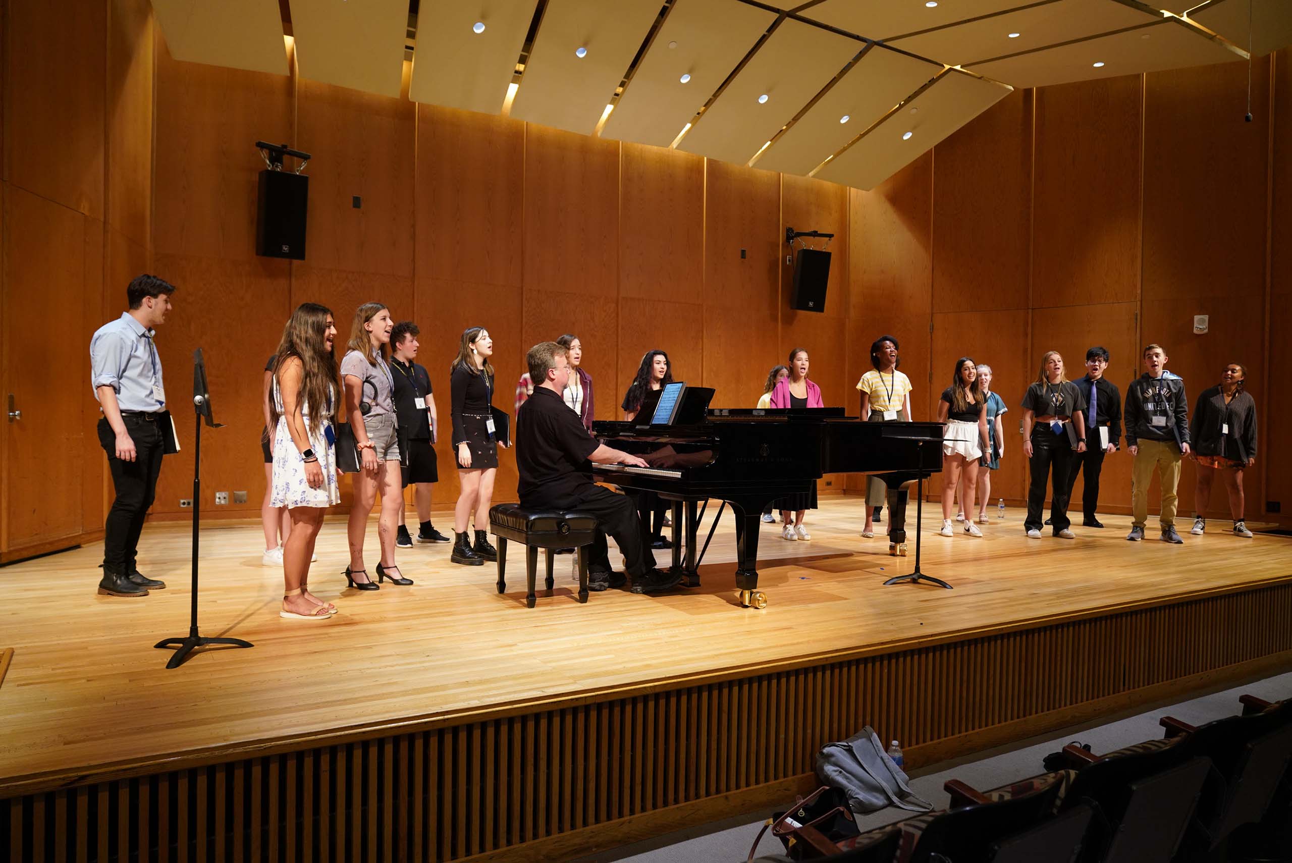 About 20 MPulse student vocalists perform standing on stage in Britton Recital Hall with a pianist.