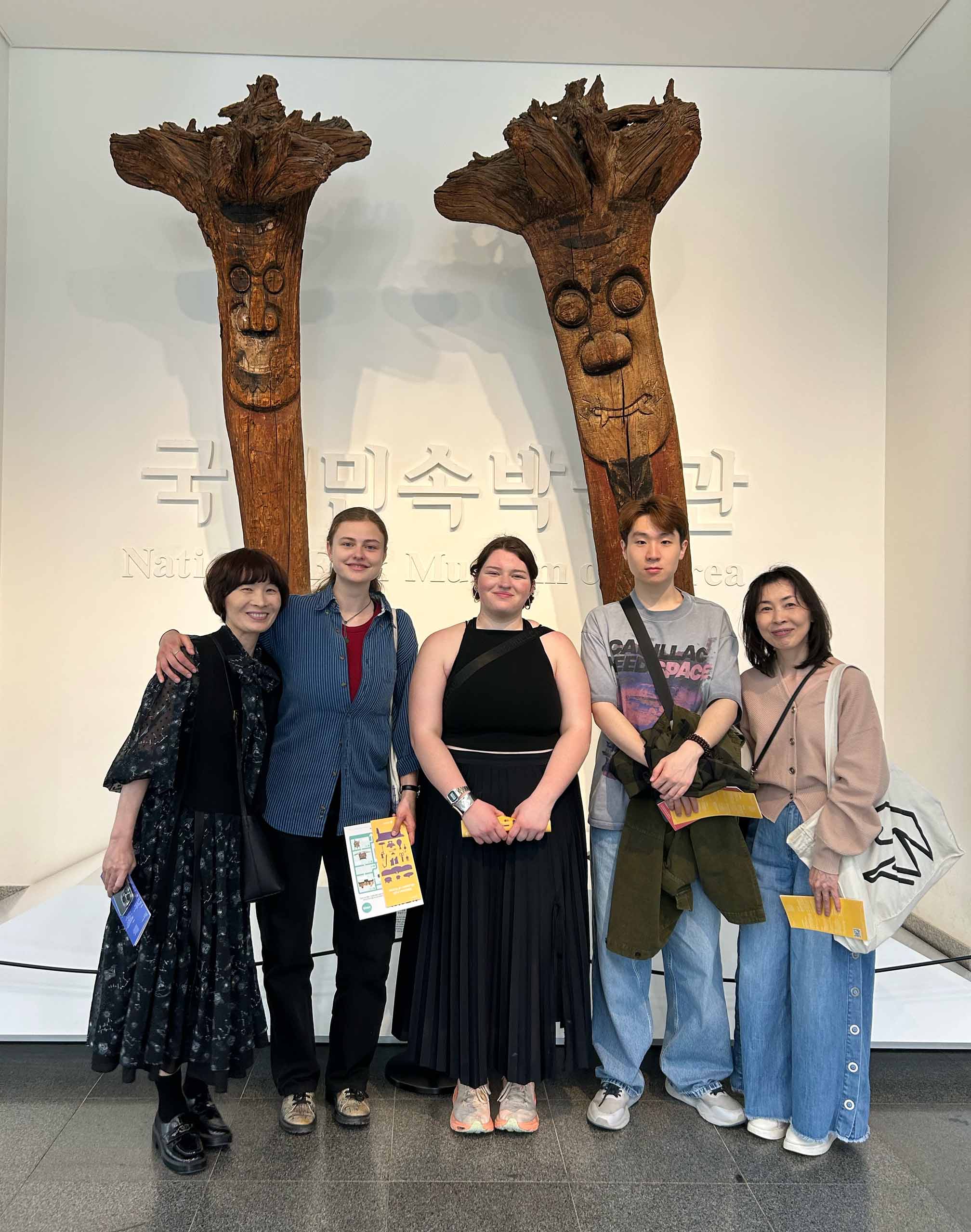 Five people pose standing together in front of an art exhibition with large Korean text on the wall.