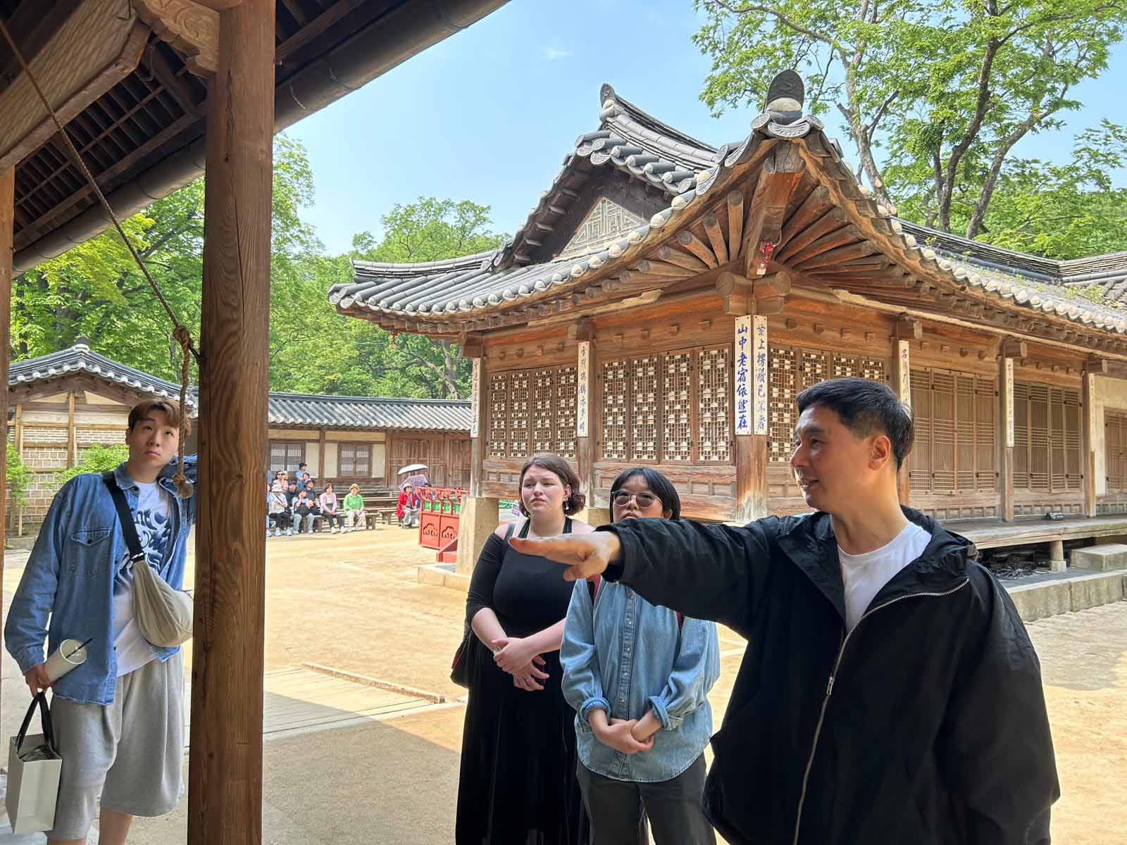 A group of four tours a historic palace grounds in Korea, as one points towards a building while speaking to the others.