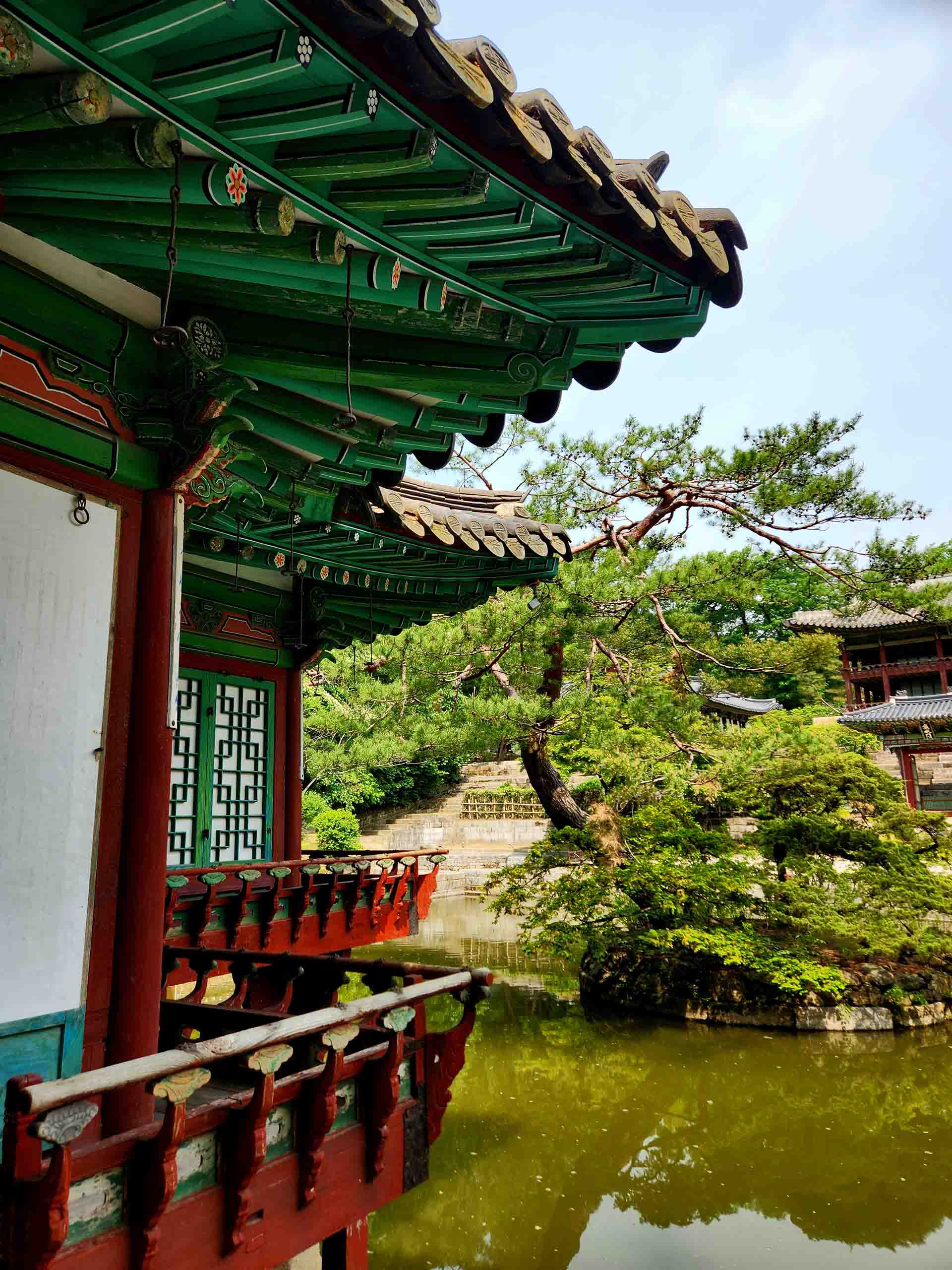 A historic Korean palace painted with red and green, with detailed architectural decoration, sits at the edge of a pond.