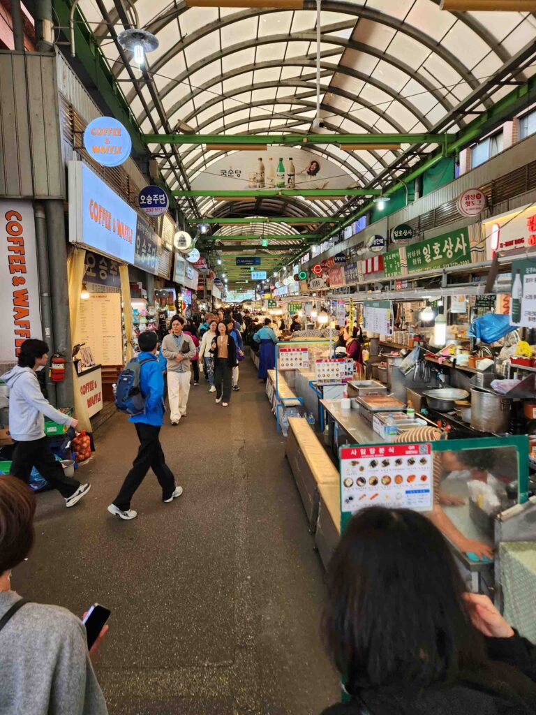 Inside a busy South Korean market with stalls of food vendors, covered by a long, slender domed ceiling.