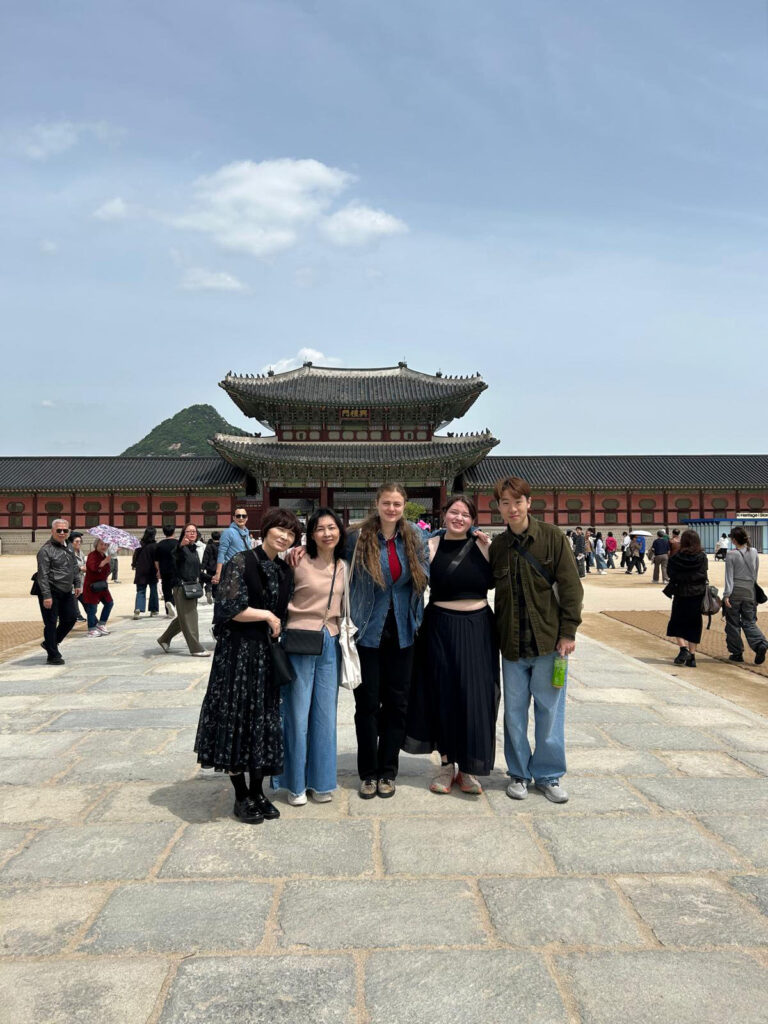 Five people pose standing in front of a historic Korean palace, on a plaza filled with many other visitors. A green mountain peak rises behind the palace in the distance.