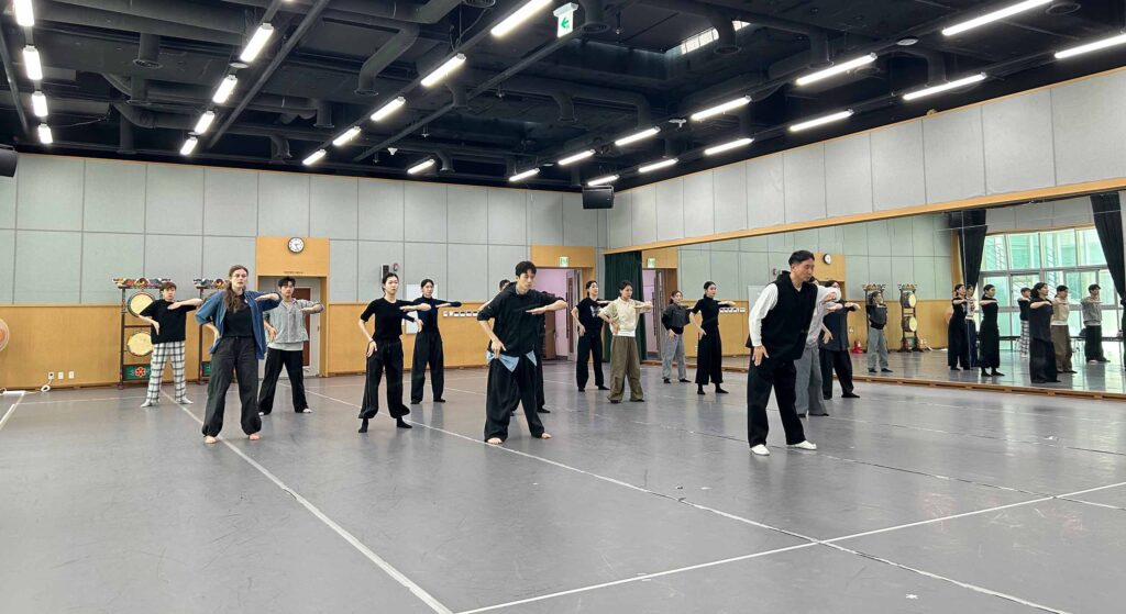 In a dance studio with a grey floor and mirror wall, about fourteen dancers stand in several rows, following a leader holding one elbow raised and a flat palm facing down.