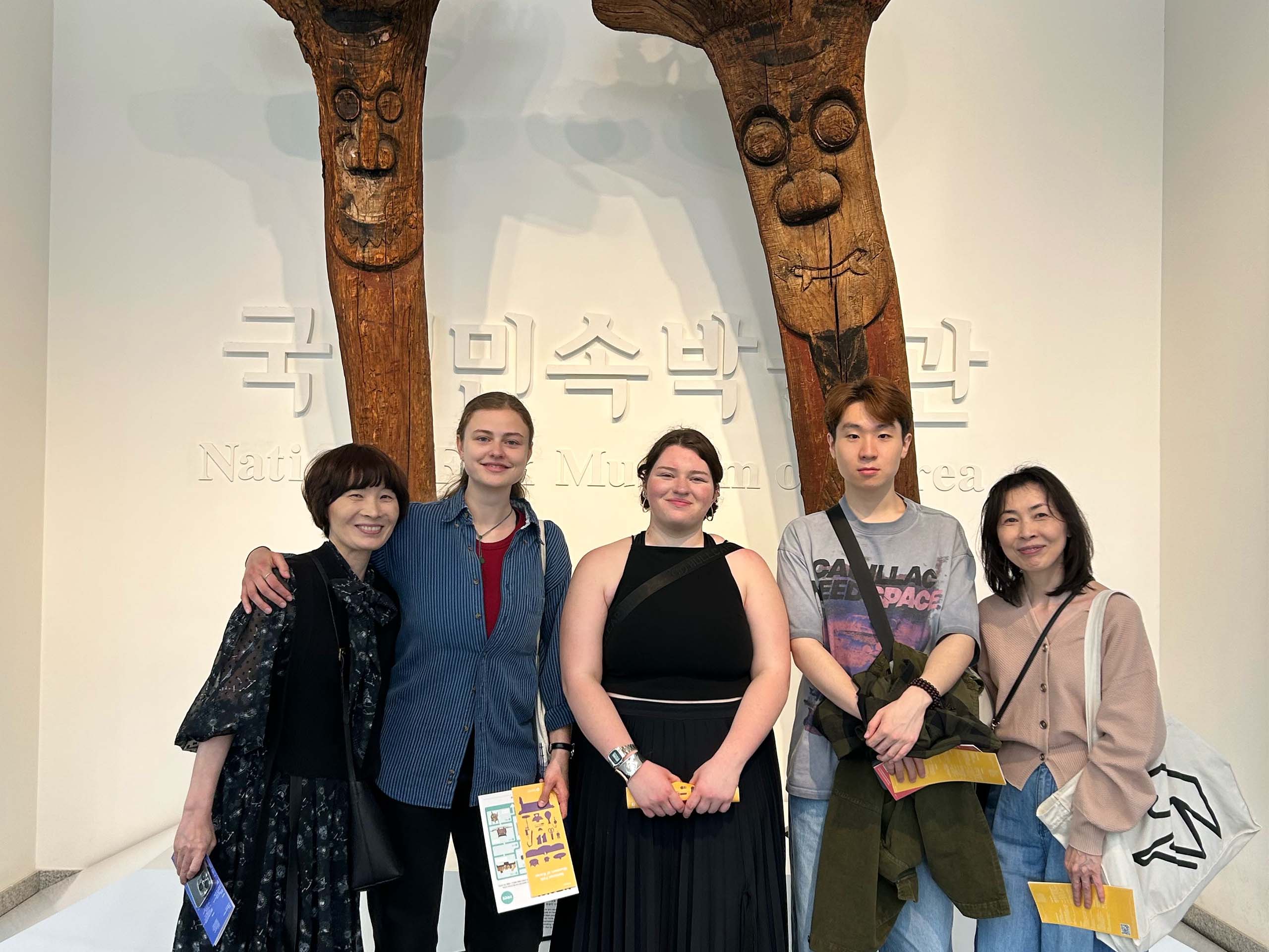 Five people pose standing together in front of an art exhibition with large Korean text on the wall.