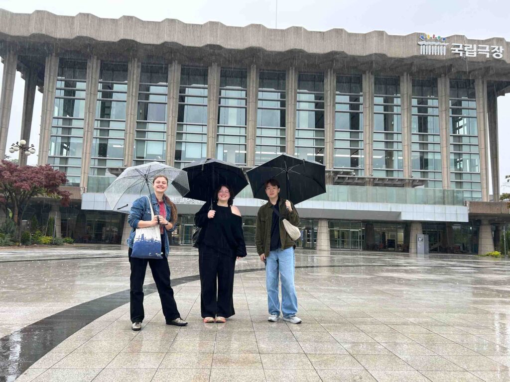 Three people pose standing in the rain, each under their own umbrella, in front of a large modern building with multi-story glass walls, and exterior concrete columns and roofline.
