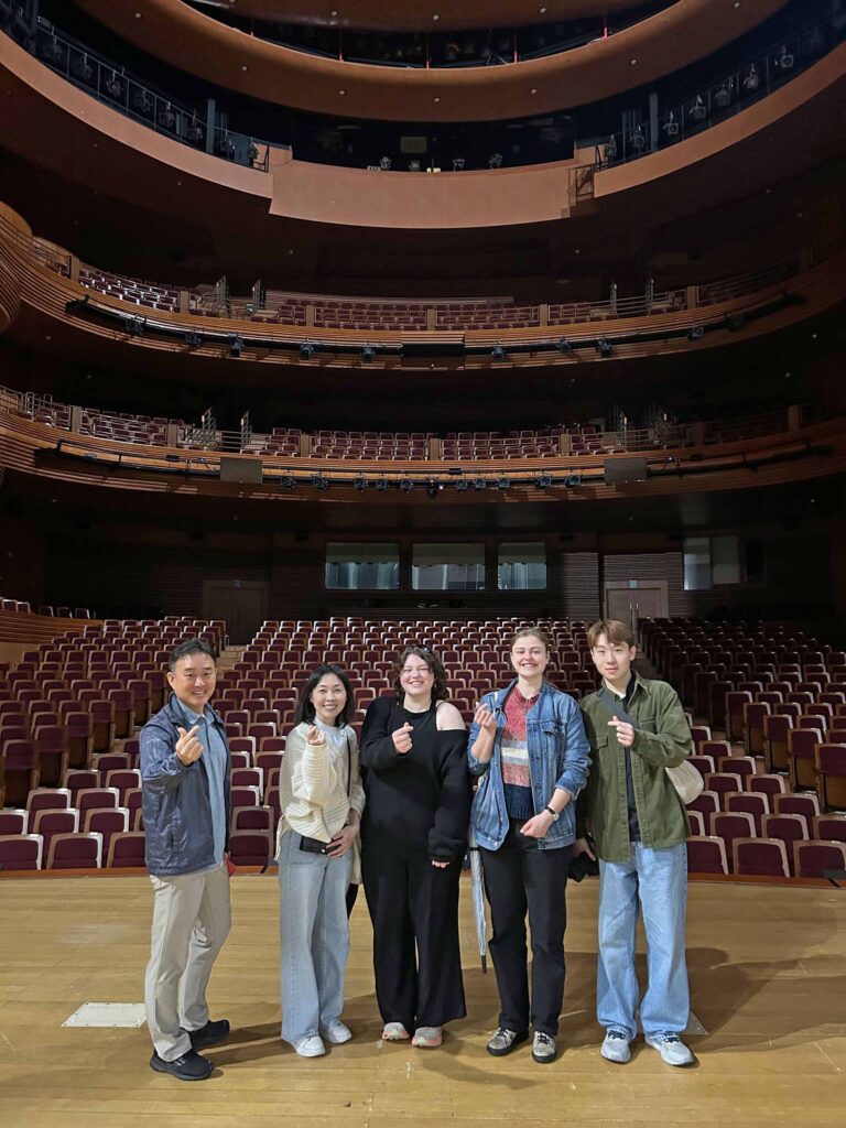 Five people pose standing on a stage making the same hand gesture. Behind them, a soaring theatre house in brown hues has a rounded mezzanine and balcony.