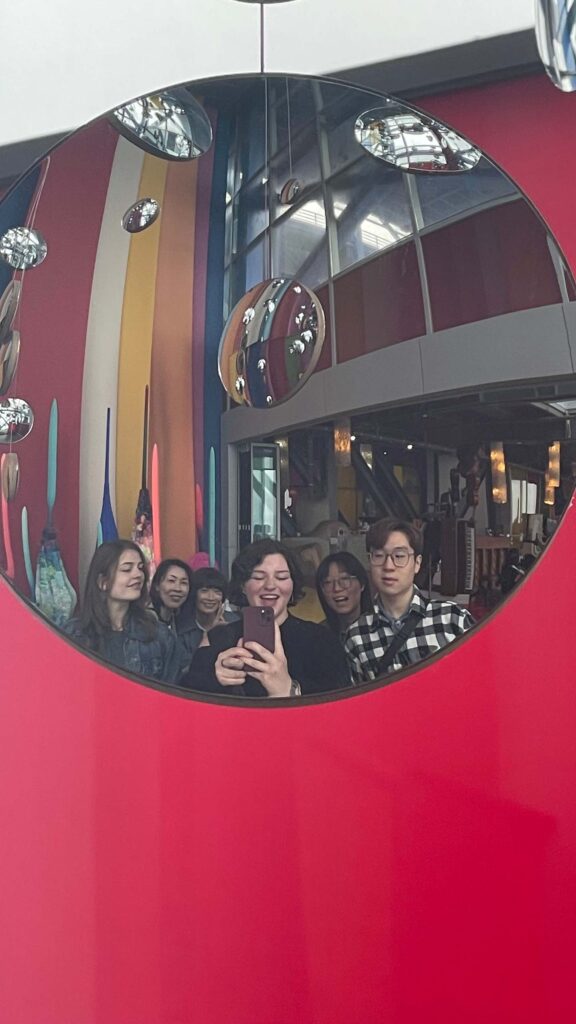 Mobile selfie taken looking into a round mirror on a red wall, with six people posing, and other round mirrors hanging in various alignments above them.