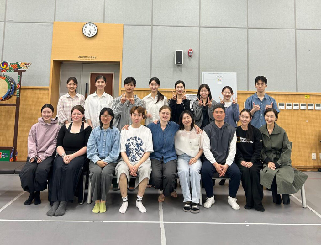 A group of 17 people pose together in two rows, a seated row with a second row standing. Some hold up two fingers with both hands.