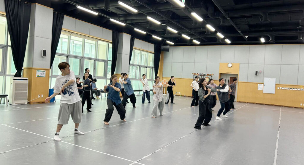 In a dance studio with a grey floor and a wall of windows, about a dozen dancers stand in synchronized motion with their elbows bent.