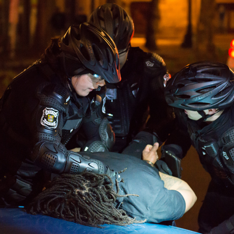 Three police officers wearing bike helmets and uniforms handcuff a person pressed face-down on a car.