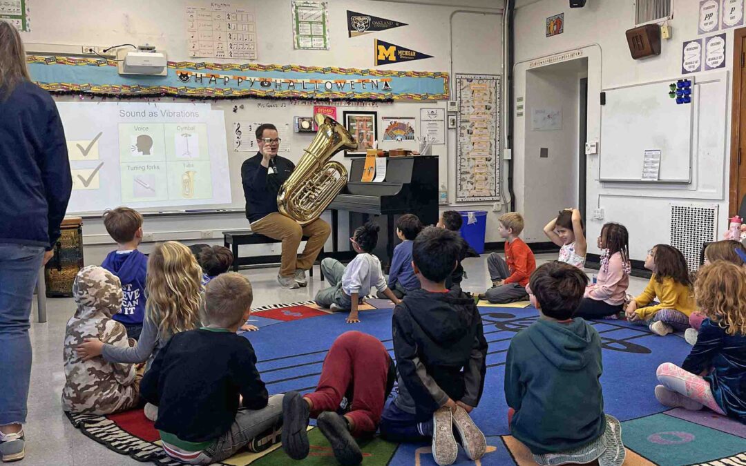 In an elementary classroom, children seated on a round carpet listen to Ryan Lofland, who holds his tuba and mouthpiece.