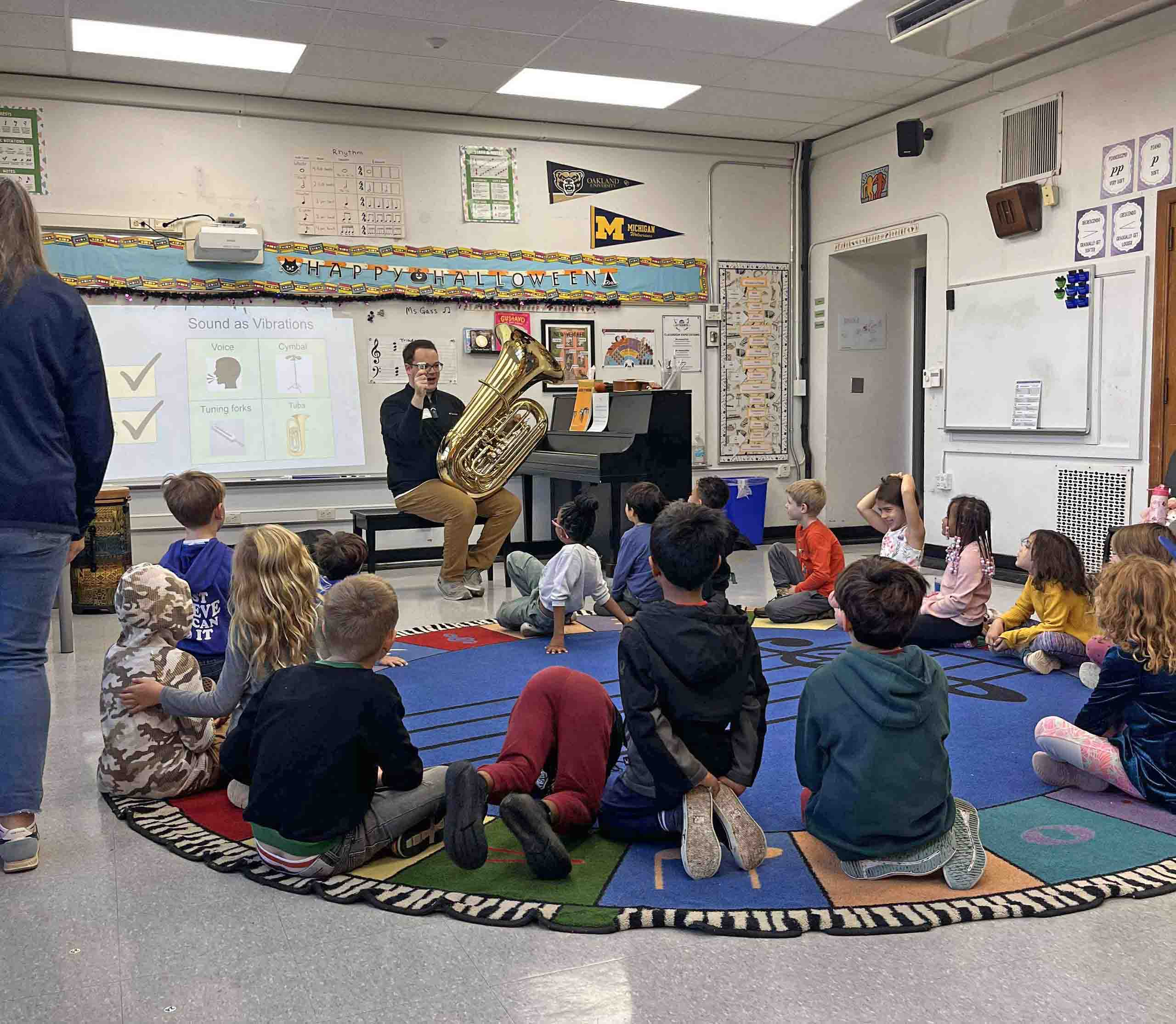 In an elementary classroom, children seated on a round carpet listen to Ryan Lofland, who holds his tuba and mouthpiece.