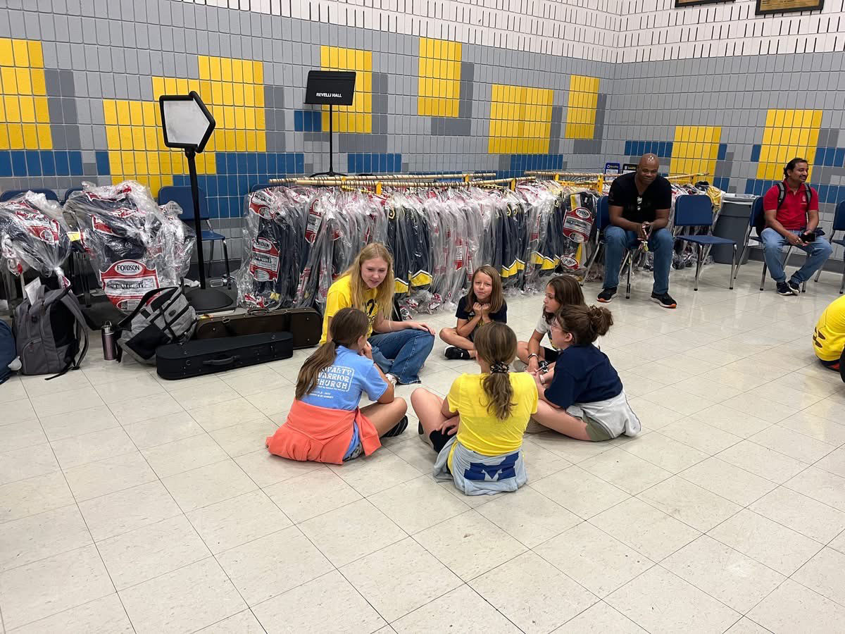 Lauren Troutman talks with five elementary school students, all seated in a circle on the floor. Behind them are marching band uniforms and equipment in storage.