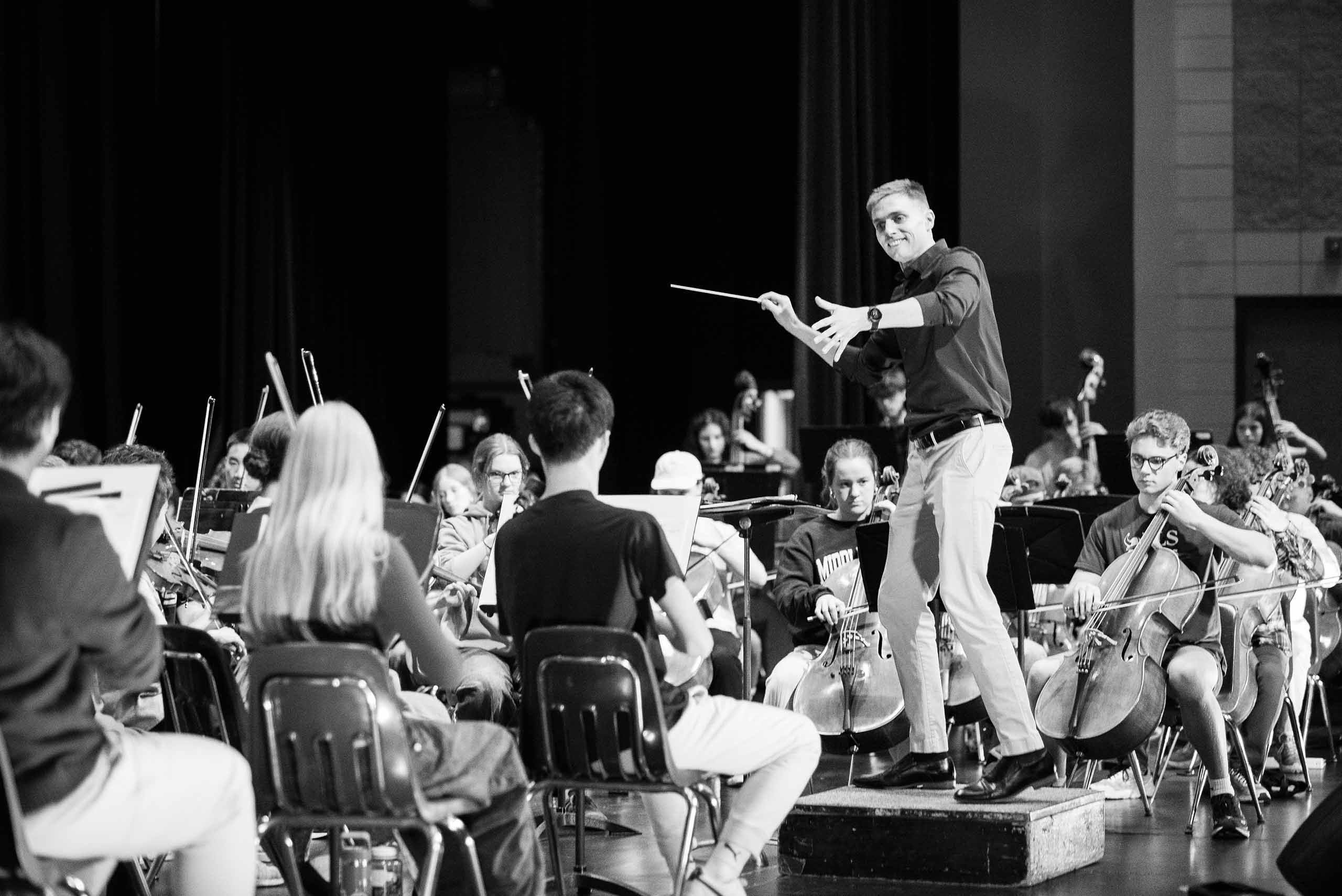Joel Schut conducts a student orchestra rehearsal in an auditorium space.