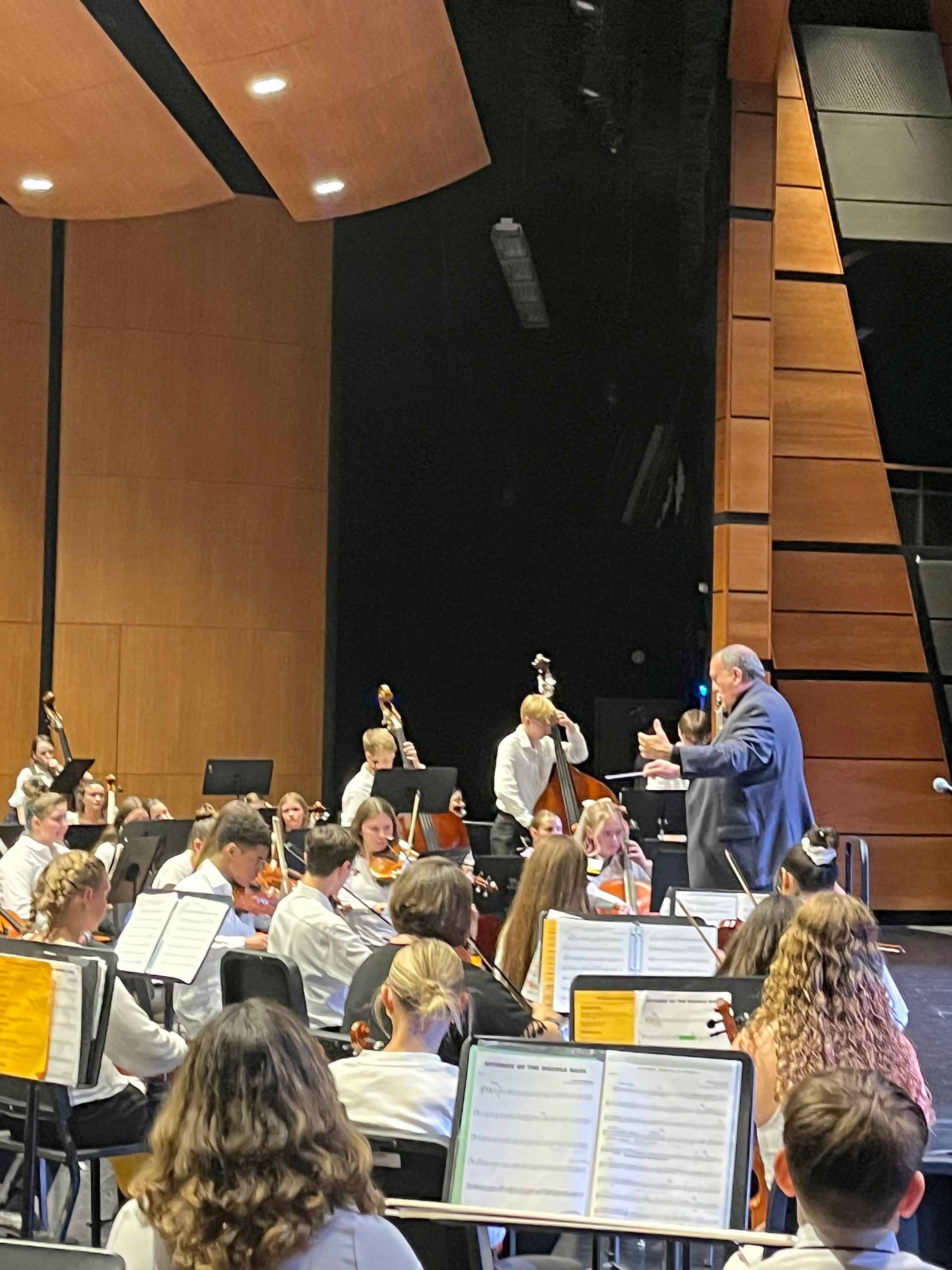 On stage in an auditorium, Bob Phillips stands conducting an orchestra of students performing dressed in white and black concert attire.