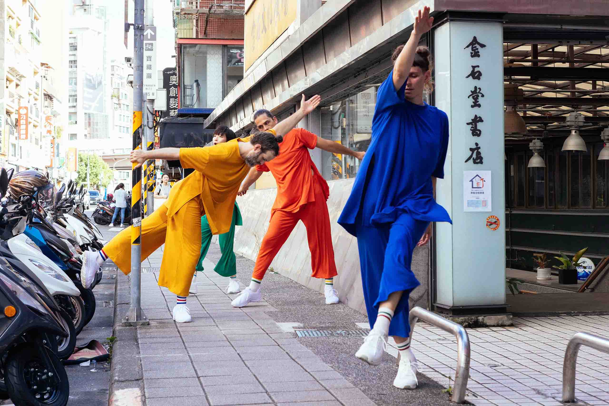 On the sidewalk of an Asian city, four performers move with long, straight arms and legs, wearing costumes of solid green, orange, yellow, and blue fabrics.