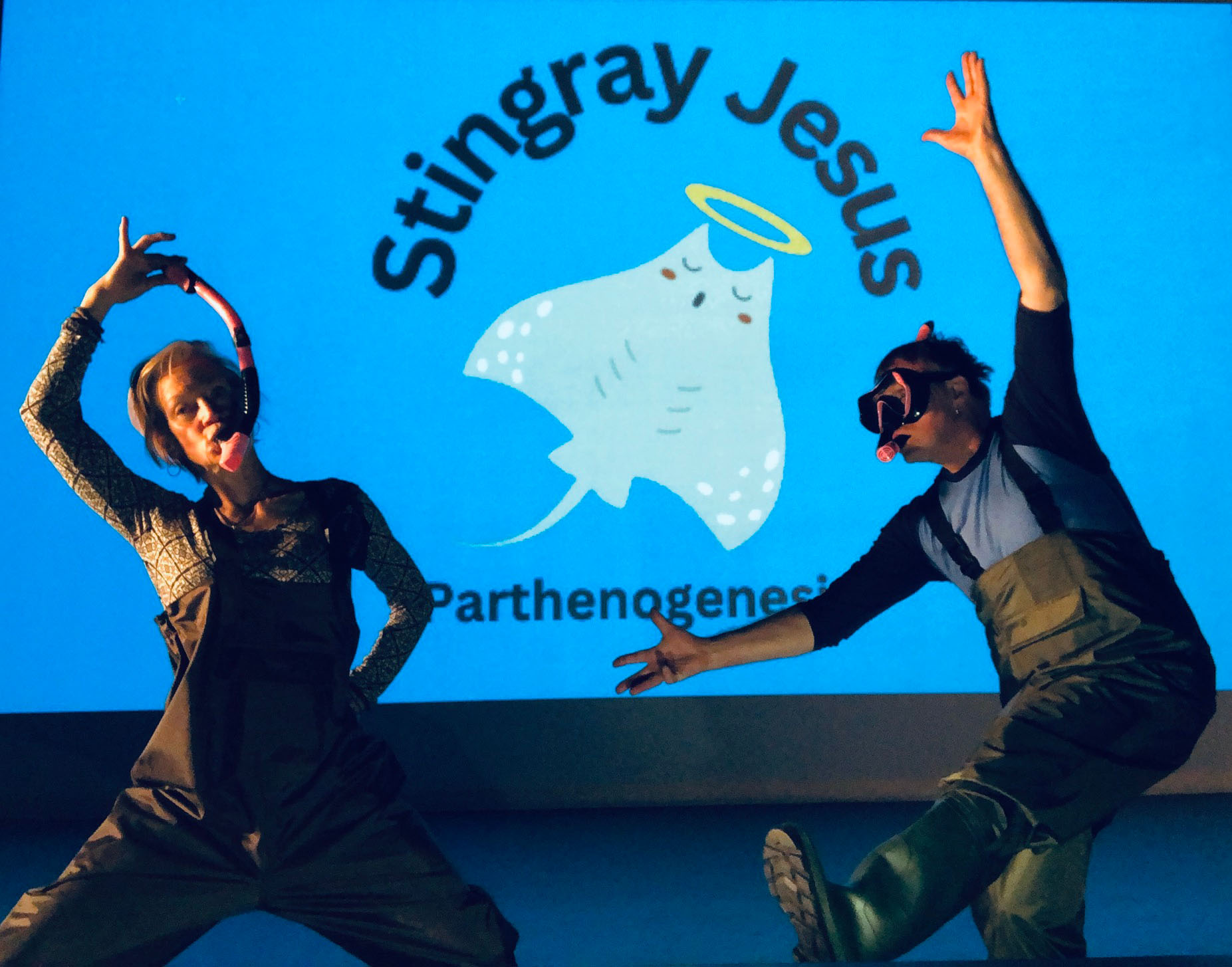 Two performers dance attired in waders and snorkeling masks, in front of a projection with a blue background that says "Stingray Jesus" with a stingray graphic.