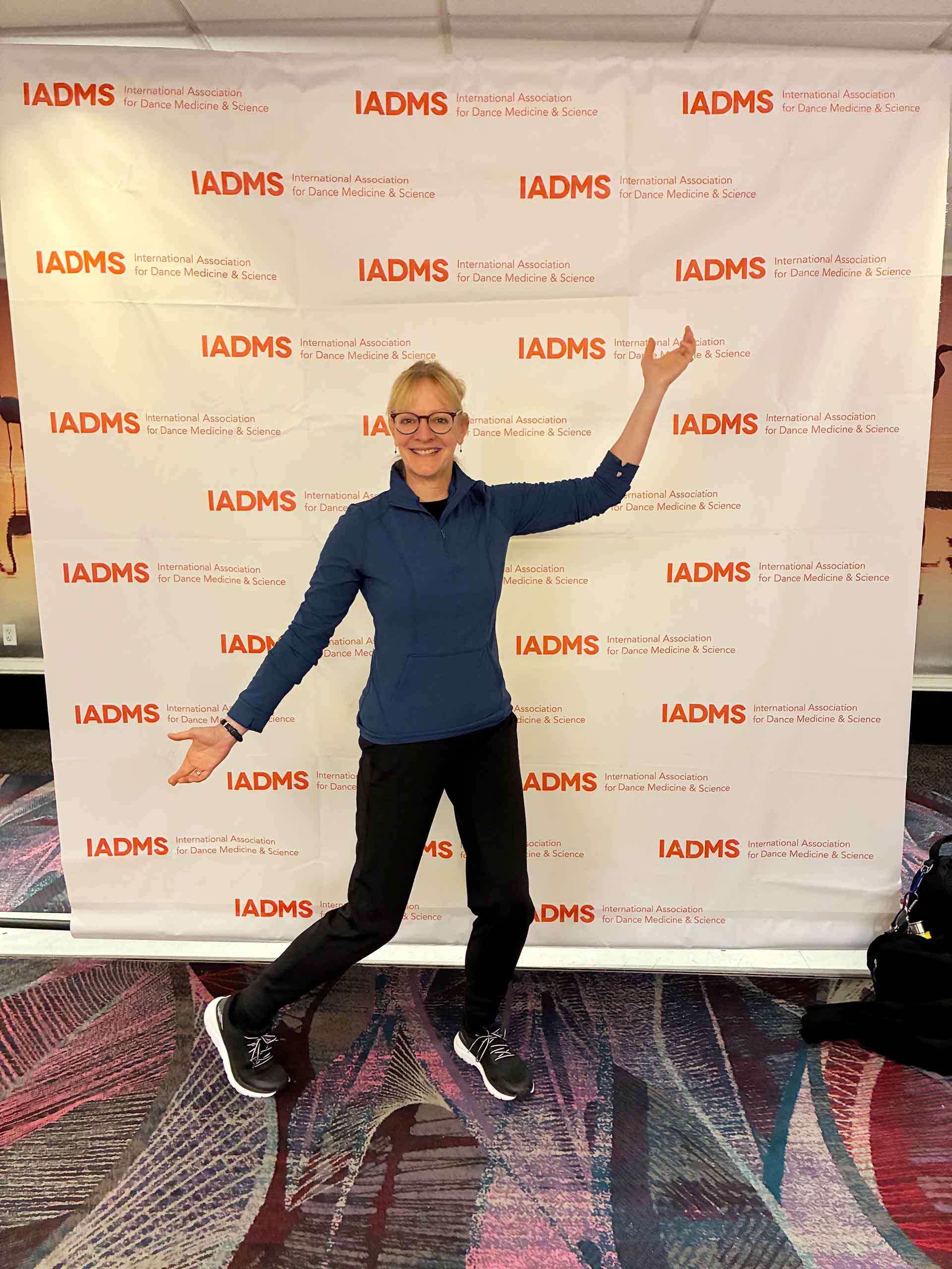 Amy West poses standing with arms outstretched in front of a conference banner.