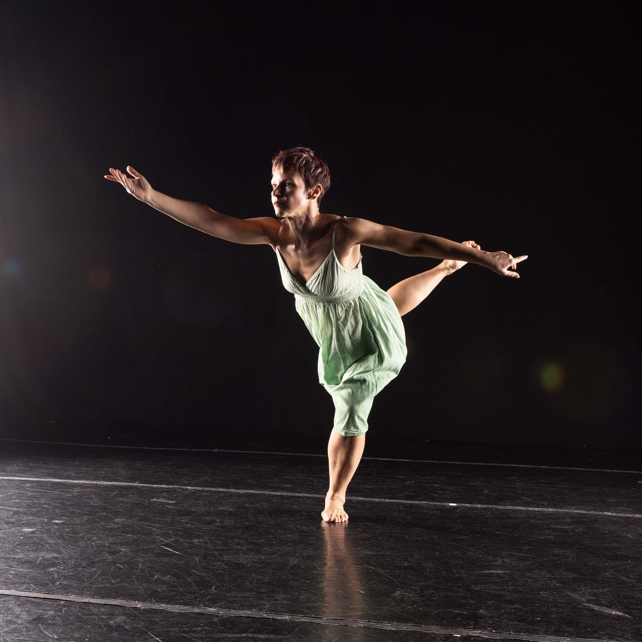 Angela Gallo holds an arabesque pose in a dark studio space.