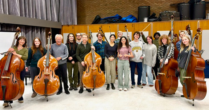A group of 16 people poses standing together in a rehearsal space, with about 6 of them holding double basses.