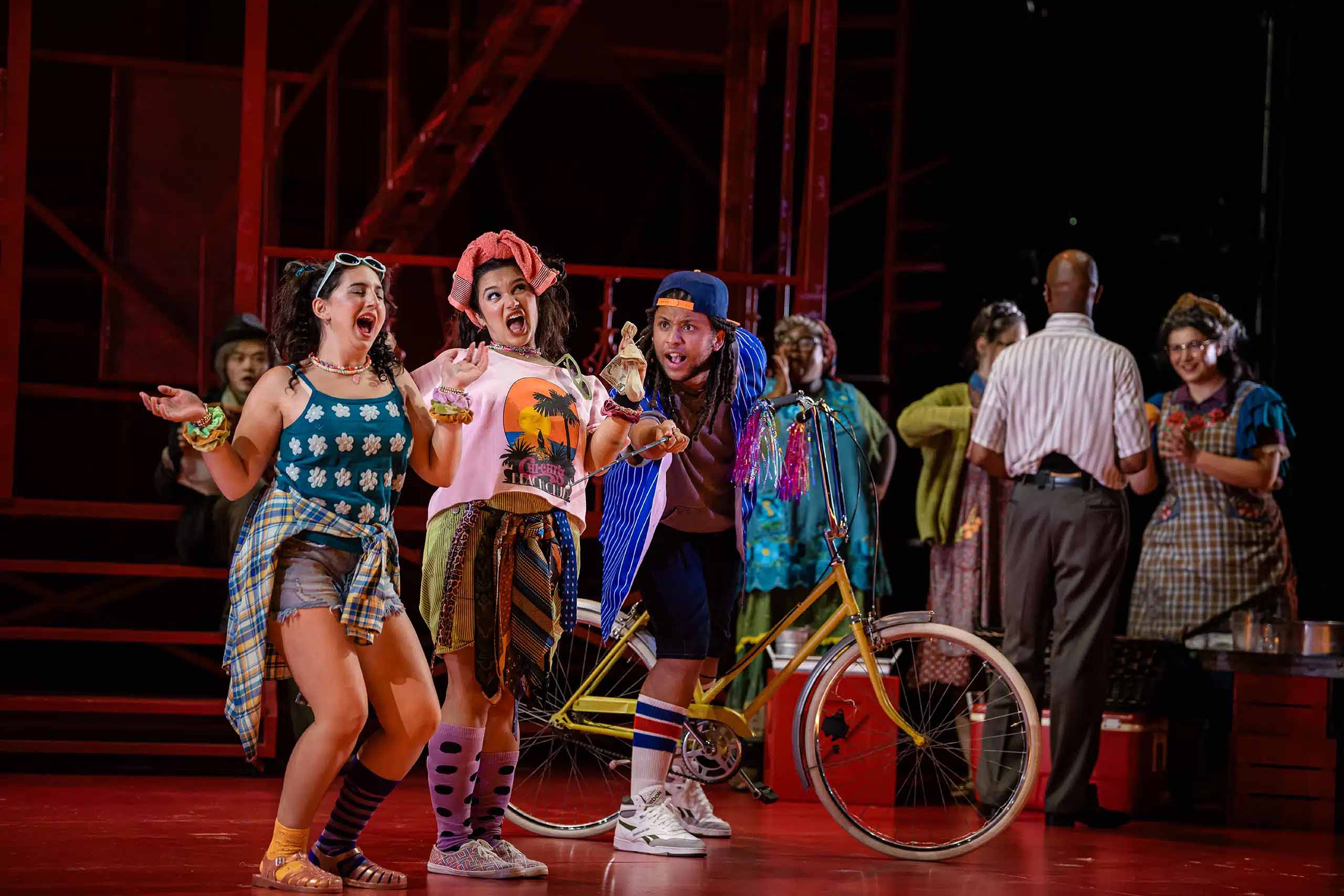 Performers sing on stage in an apparent street scene, attired in colorful and casual late 20th century costumes. One has a bike.