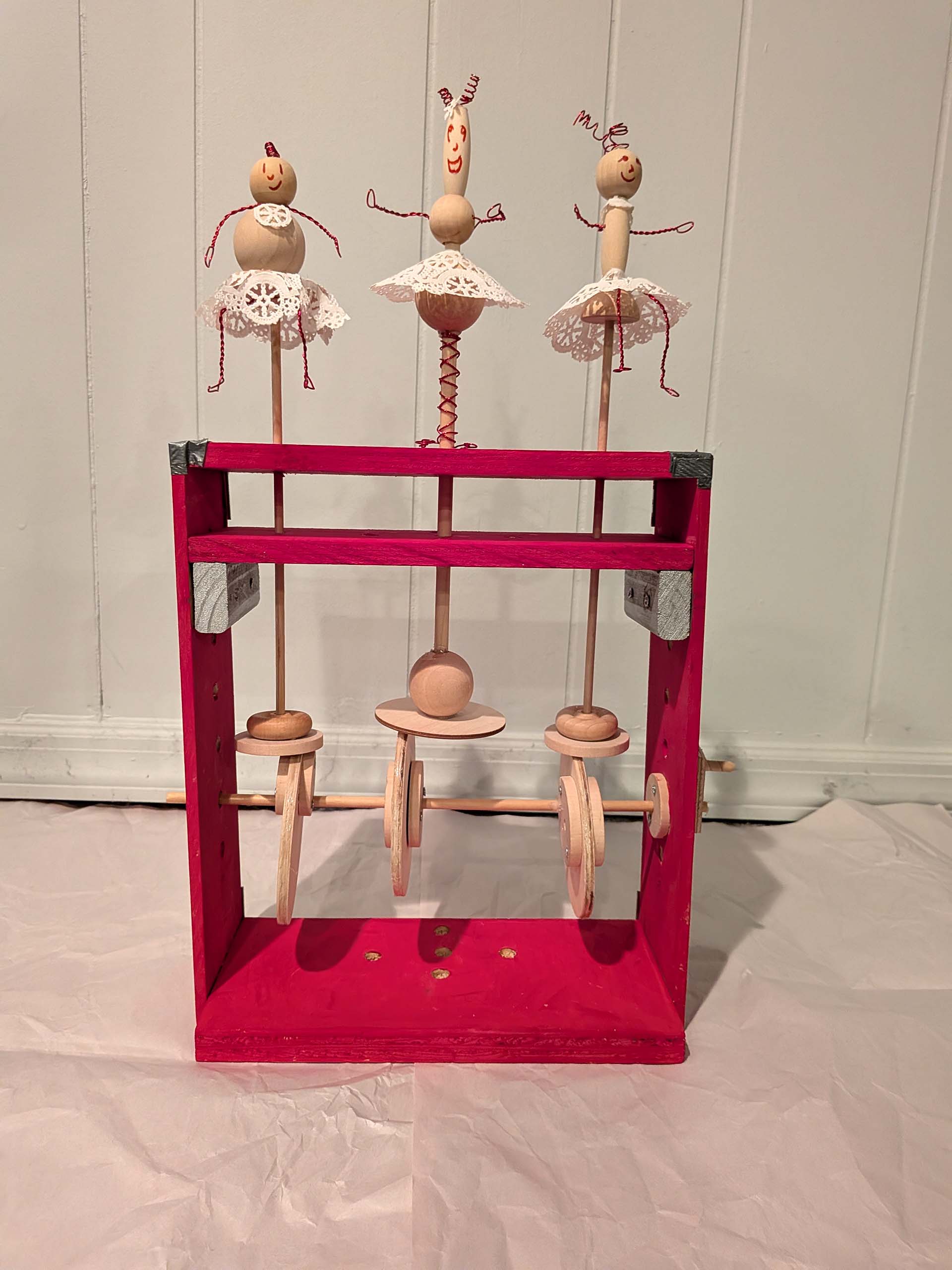 Three puppets made of wooden balls with doily skirts are attached to controls housed within a wooden frame box painted red.