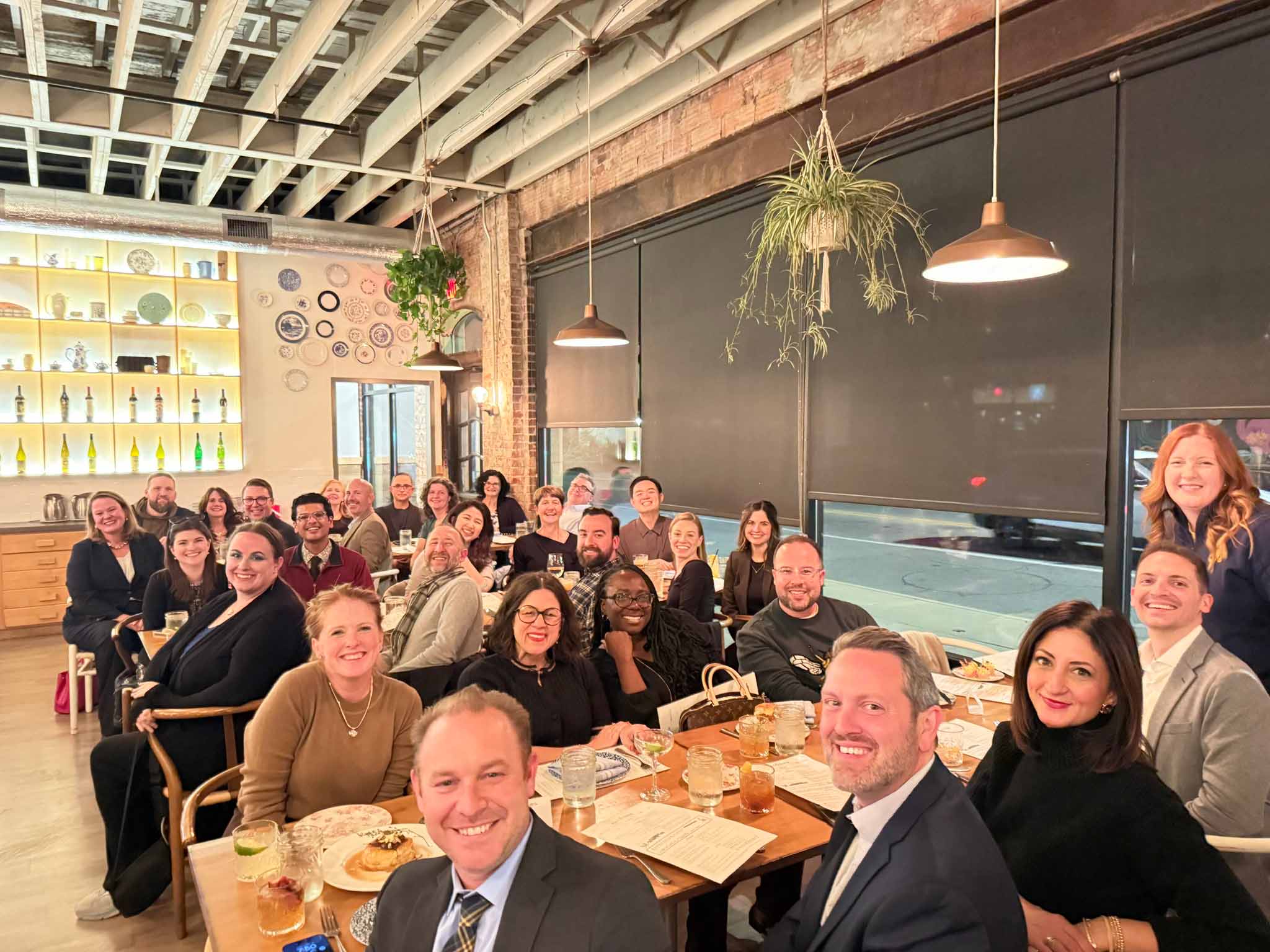 About two dozen people smile for the camera, seated at long adjacent tables while dining together at a restaurant.